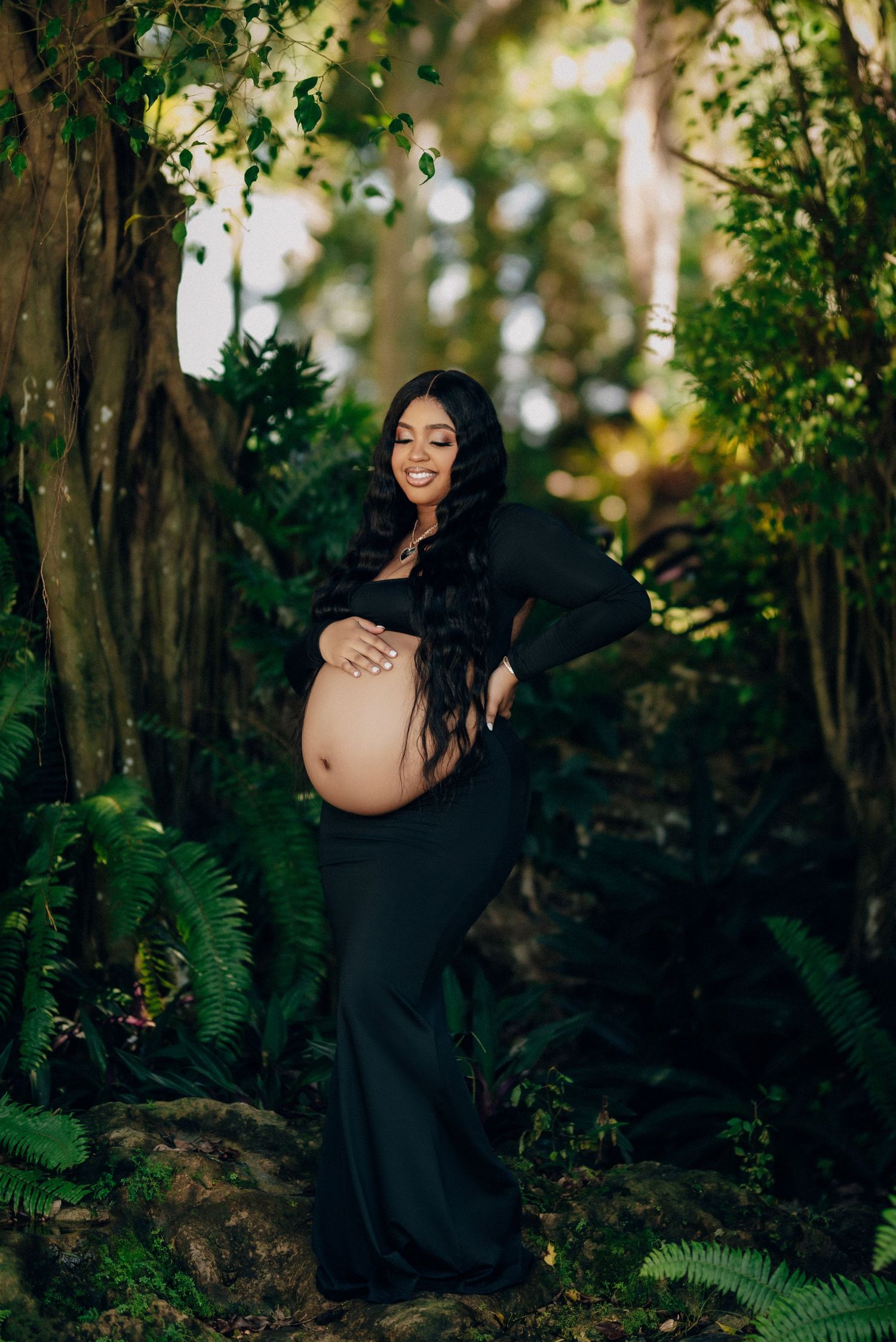 A maternity photoshoot in a lush forest setting features a woman in a flowing black dress among green foliage.