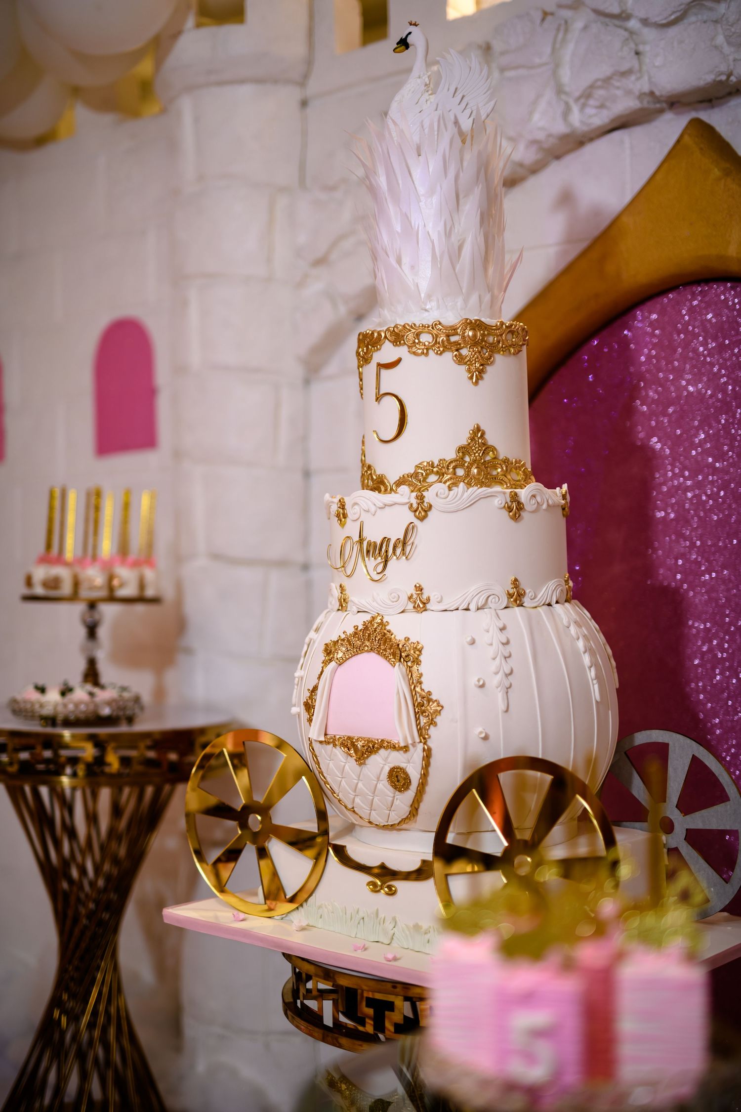 Elegant white and gold carriage-shaped wedding cake with feather topper displayed against white brick wall with pink accents.