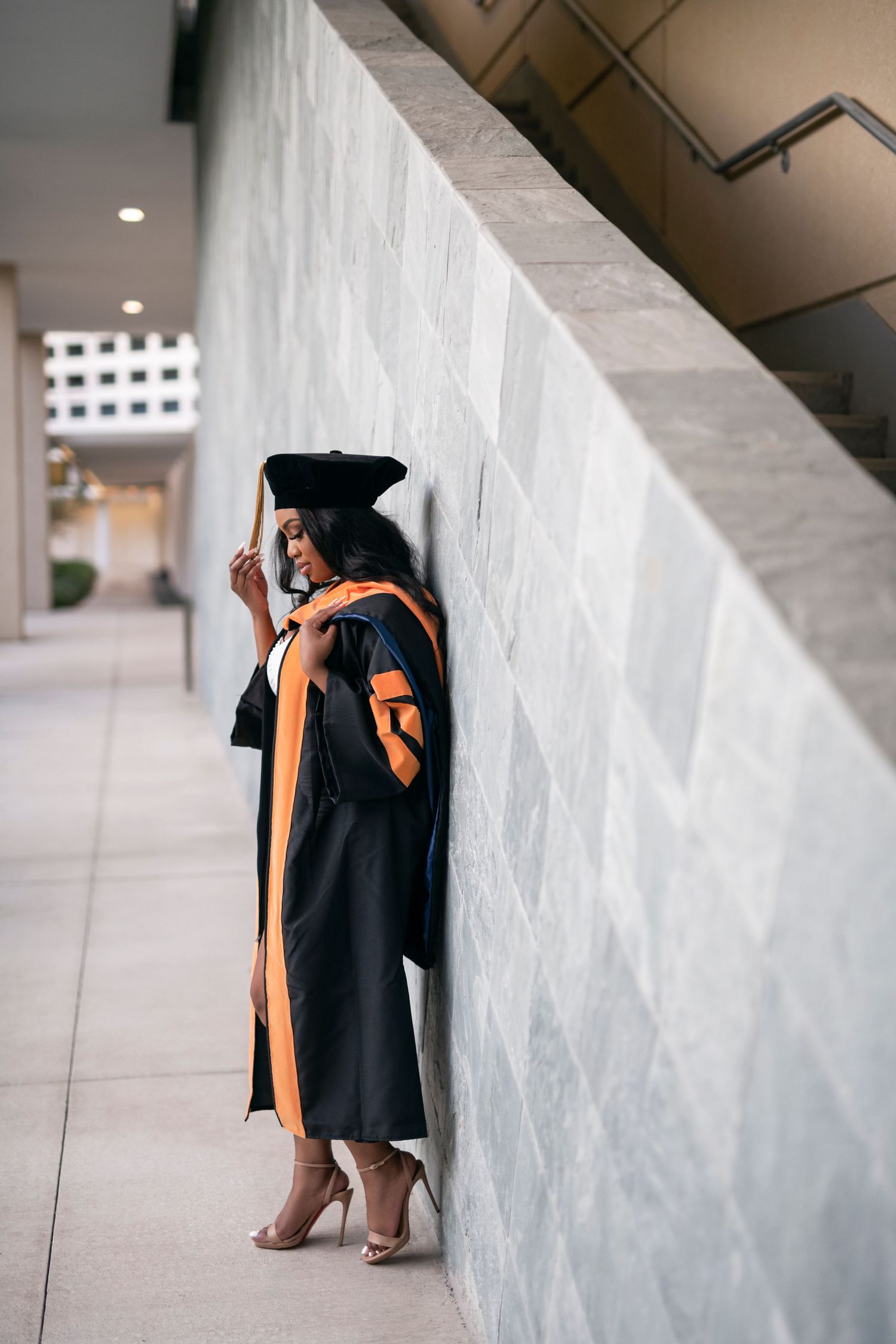 Graduate in academic regalia takes creative portraits against a modern architectural wall in a hallway.