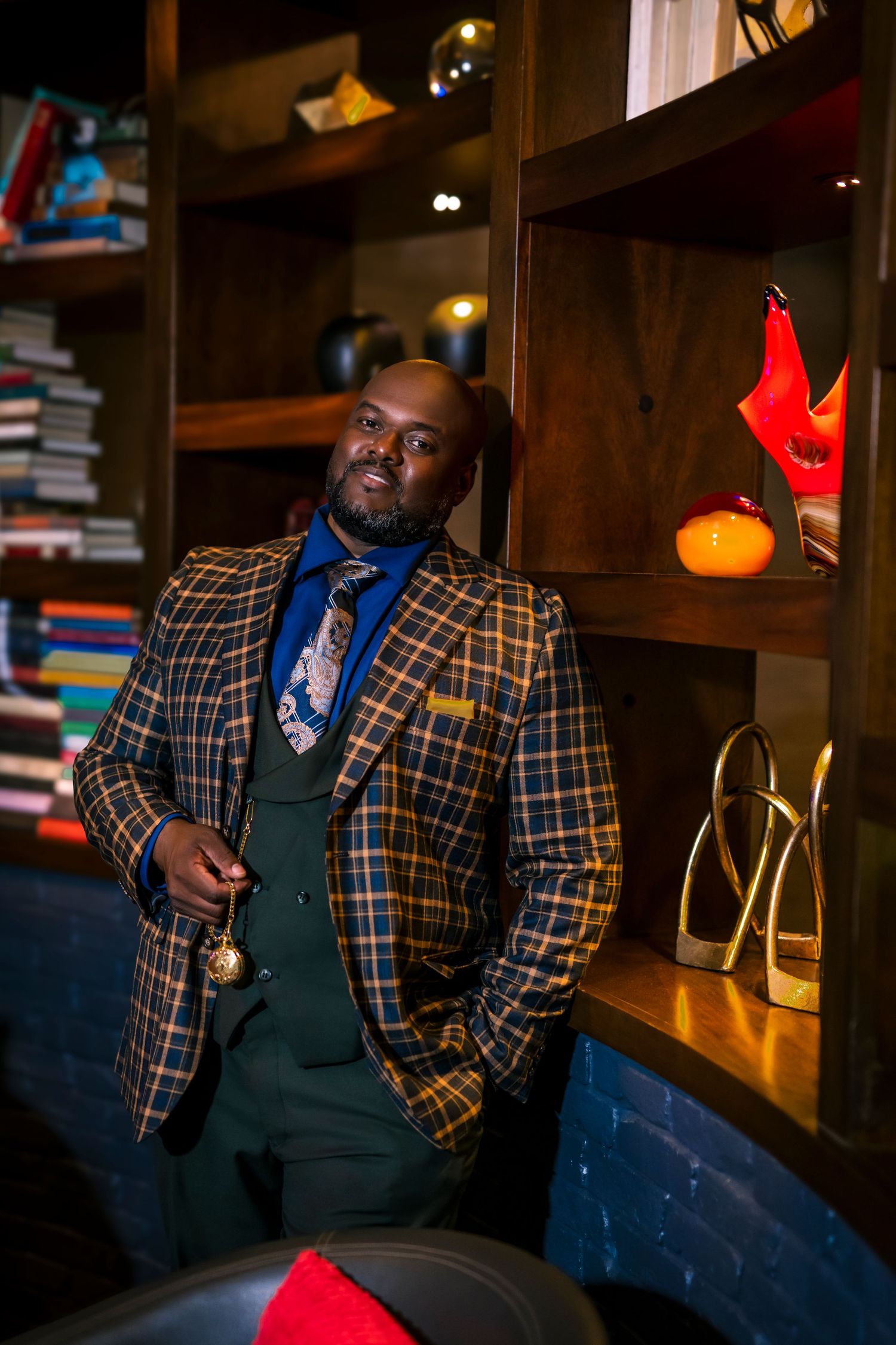 Person wearing plaid blazer and dark vest poses in moody lighting with bookshelves and decorative pumpkins.