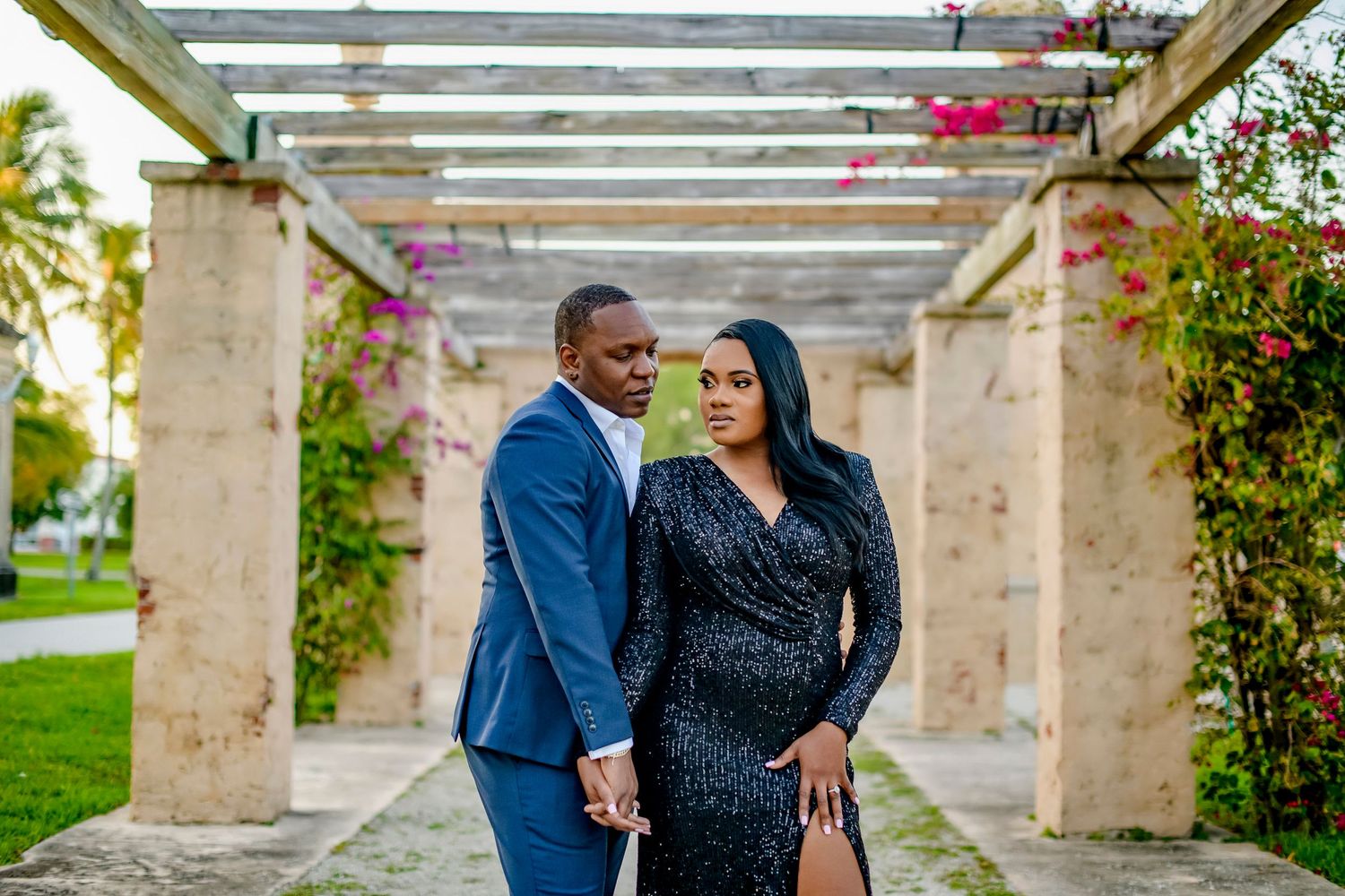A couple poses together under a garden pergola with blooming flowers, dressed in formal navy suit and sparkly black dress.