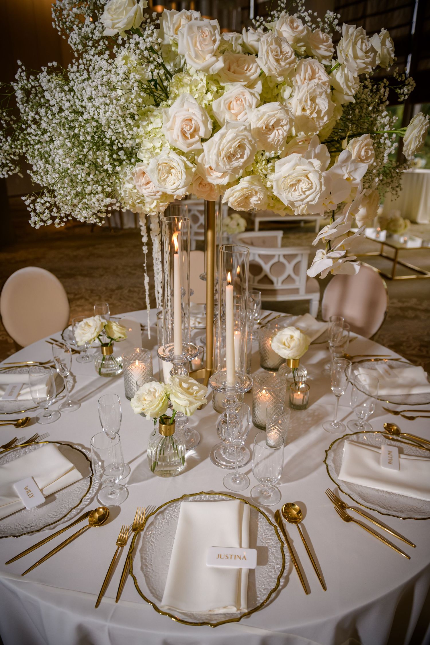 Elegant wedding reception table setting with white roses, crystal glassware, and gold accents on a round table.