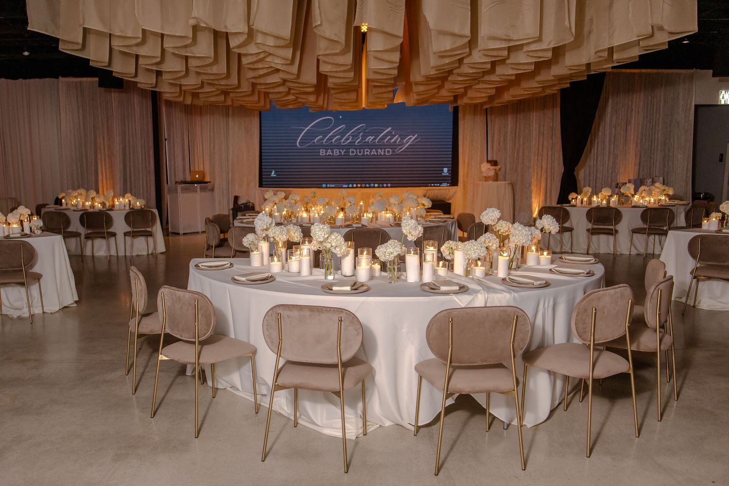 Elegant wedding reception venue with round tables, beige chairs, floral centerpieces and modern ceiling decor.