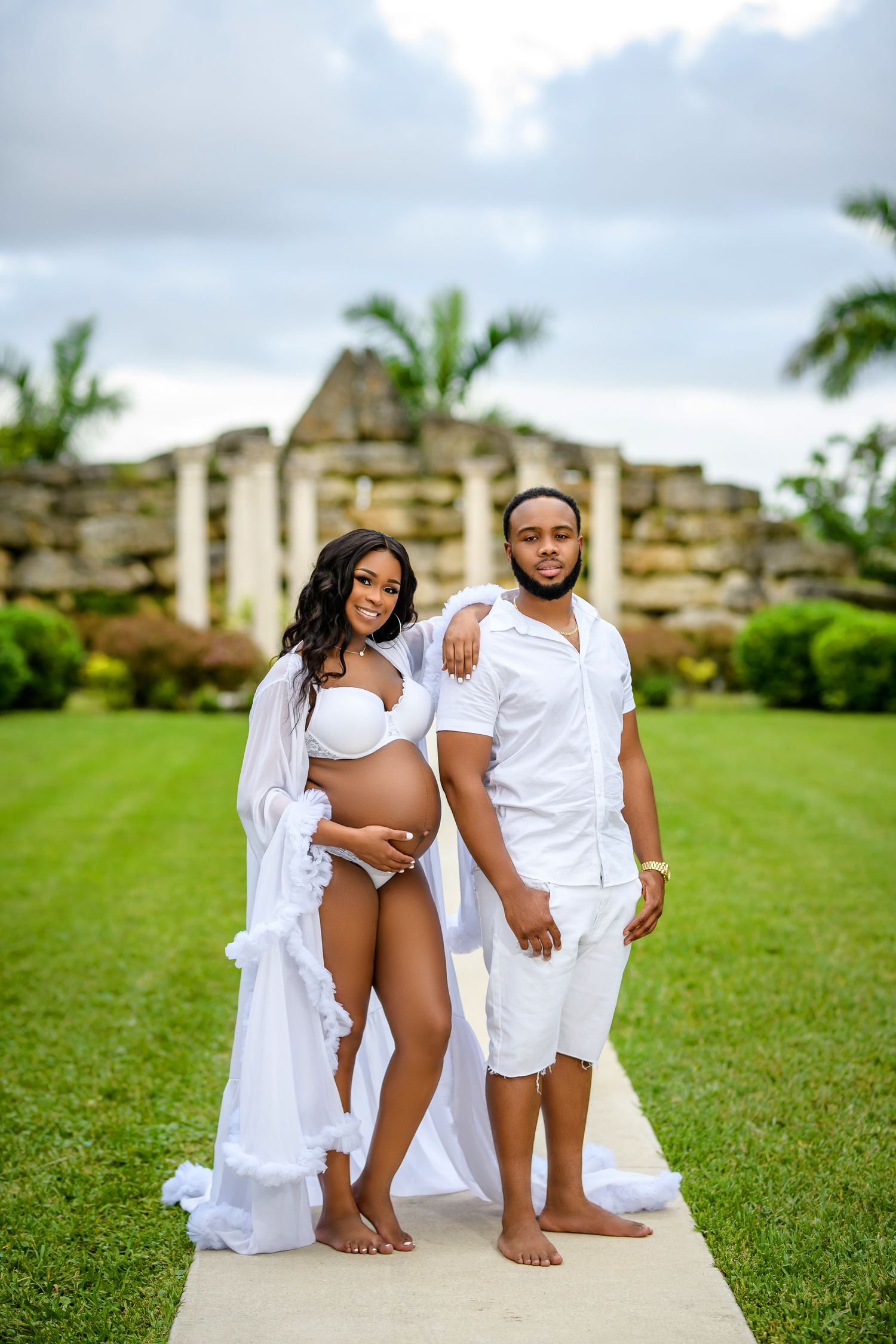 A series of artistic maternity photos taken outdoors showing a couple in white clothing on grassy grounds near stone ruins.