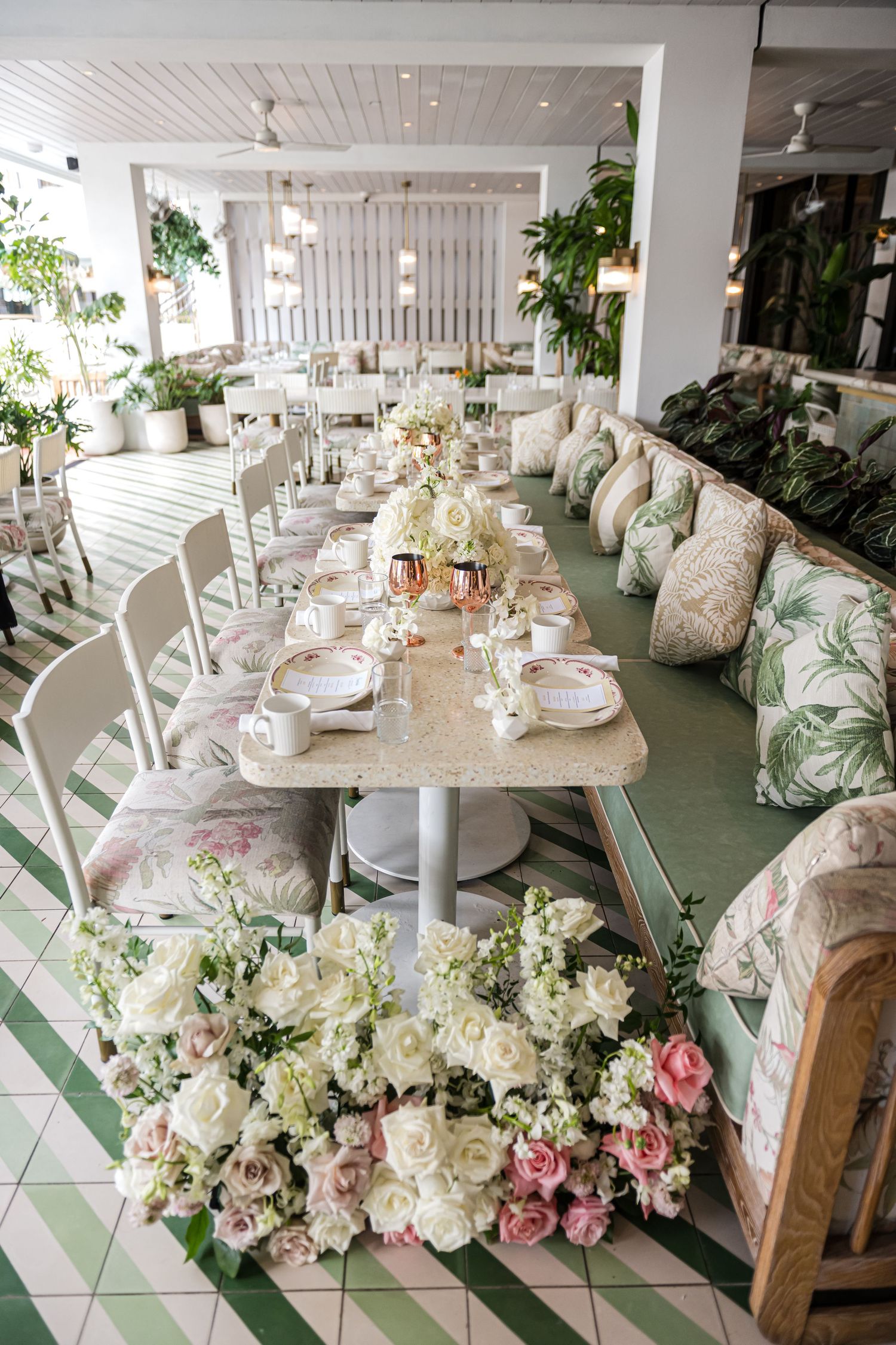Elegant restaurant interior with white tables, floral cushioned seating, and tropical leaf wallpaper decor.