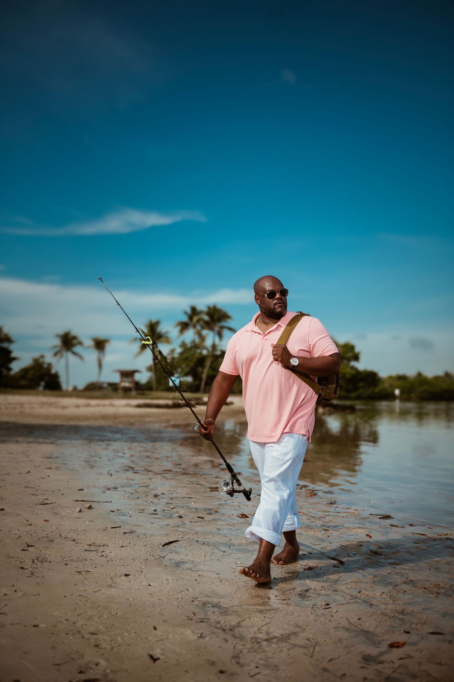 Person in pink shirt and white shorts fishing on a tropical beach with palm trees in the background.