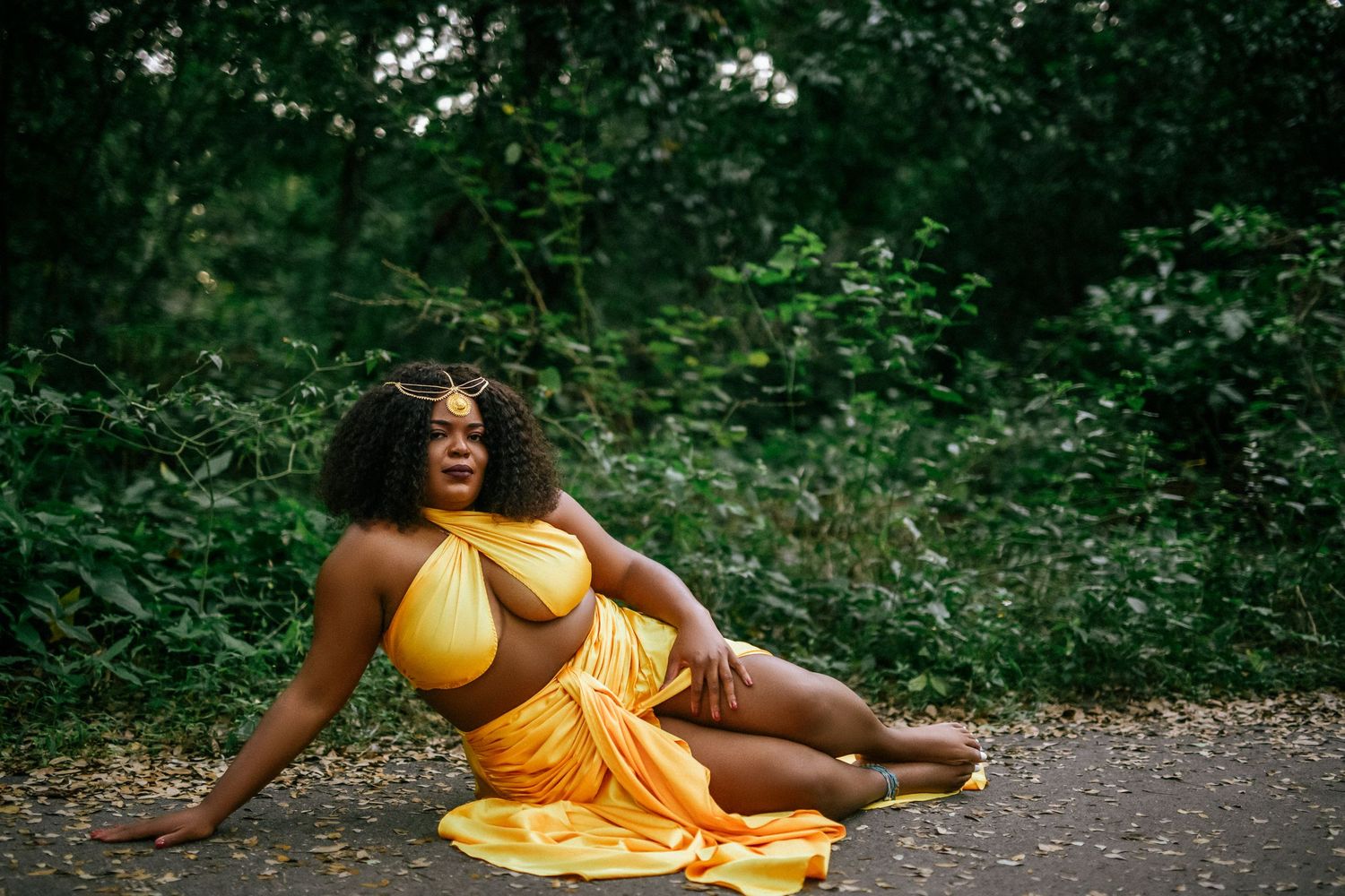 A person in a flowing yellow dress reclines on a path surrounded by lush greenery.