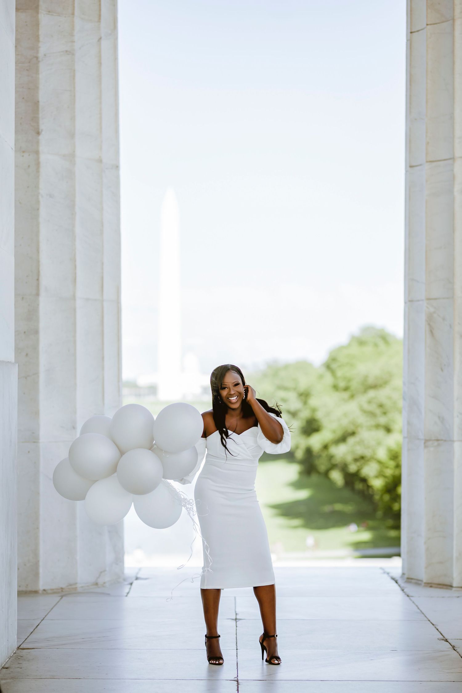 A woman in a white dress poses with white balloons between marble columns on a bright sunny day.
