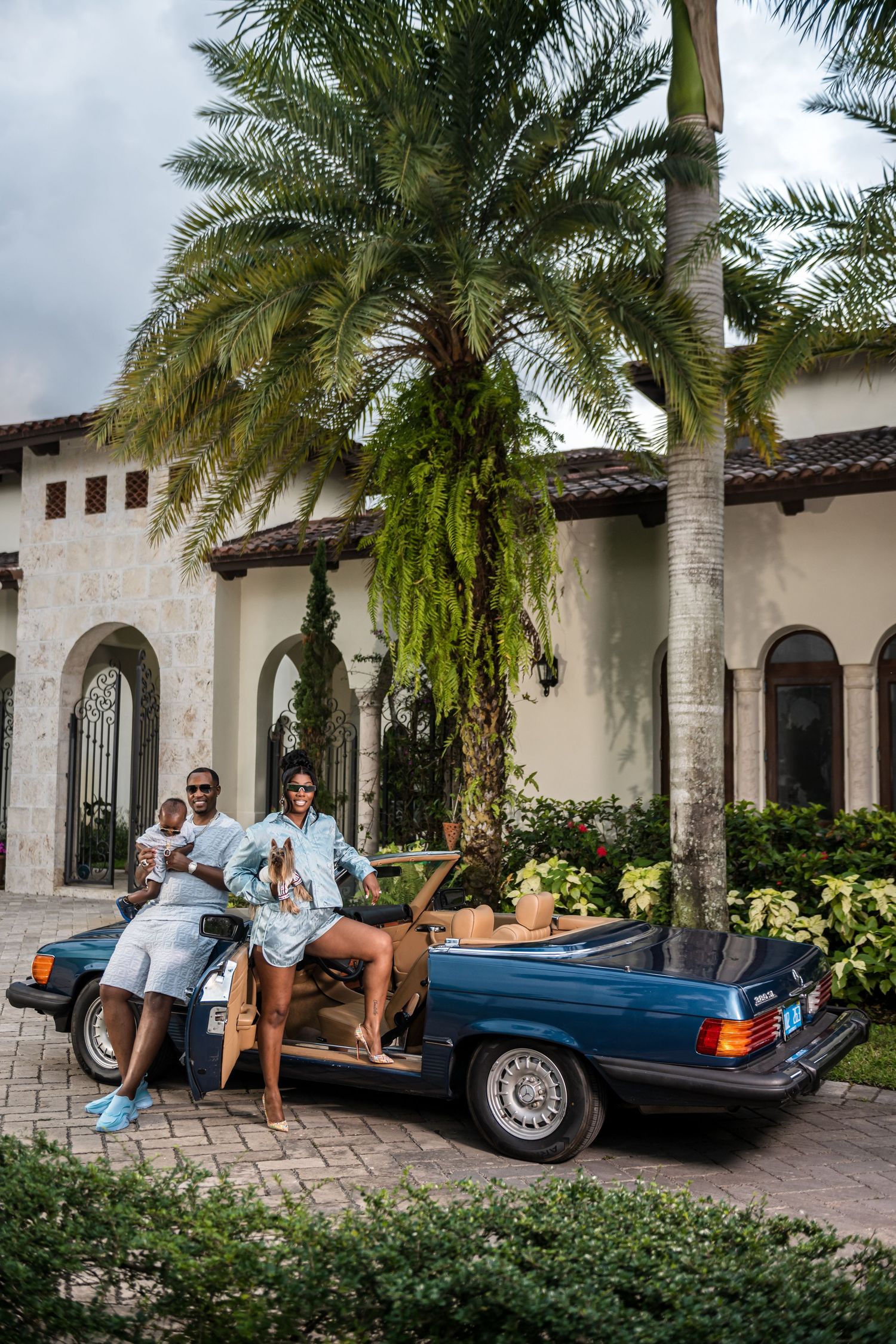 People posing with vintage blue convertible car in front of Mediterranean style villa with palm trees.