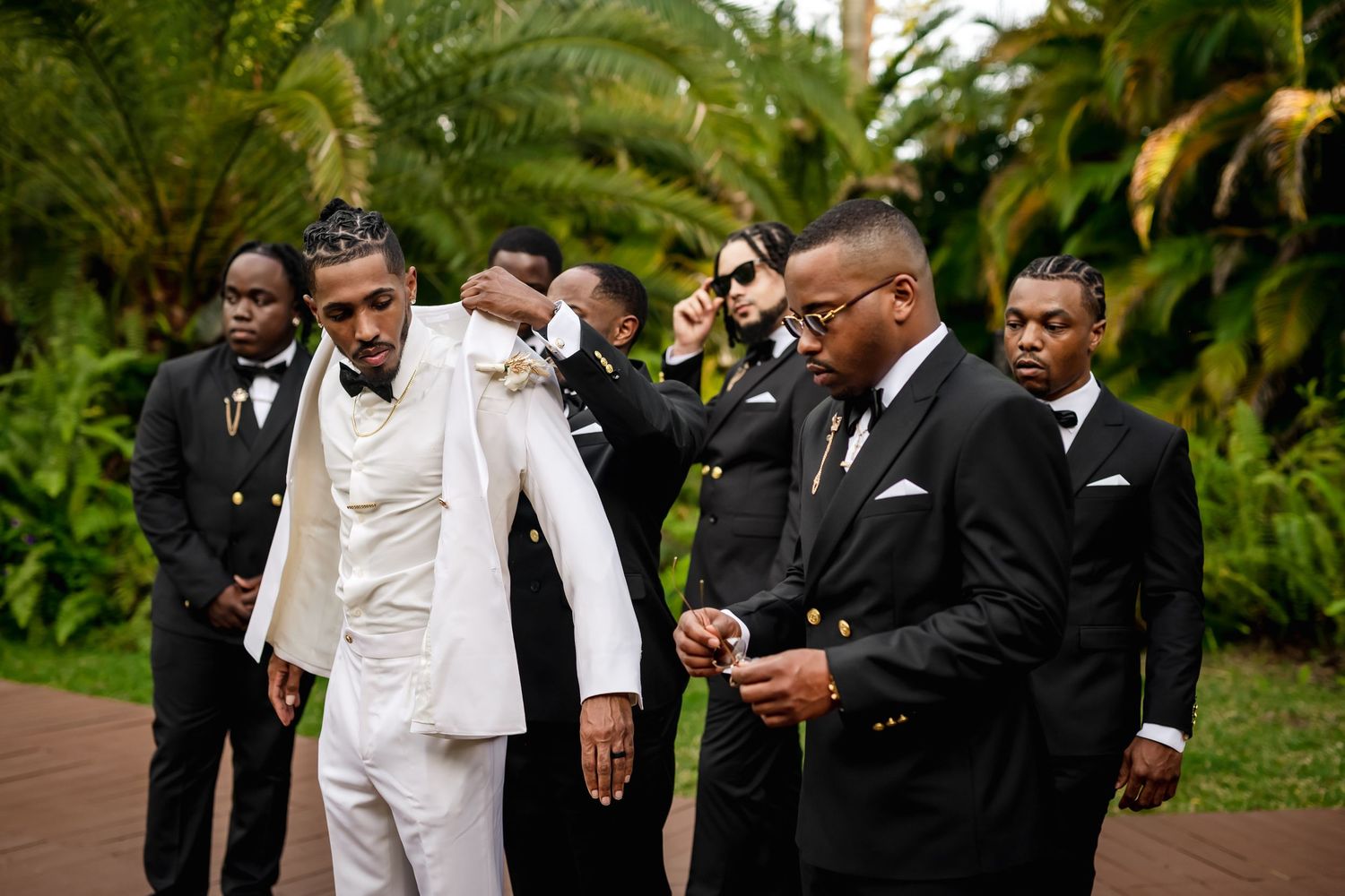 A group of groomsmen in black and white formal attire gather together in a lush tropical garden setting.