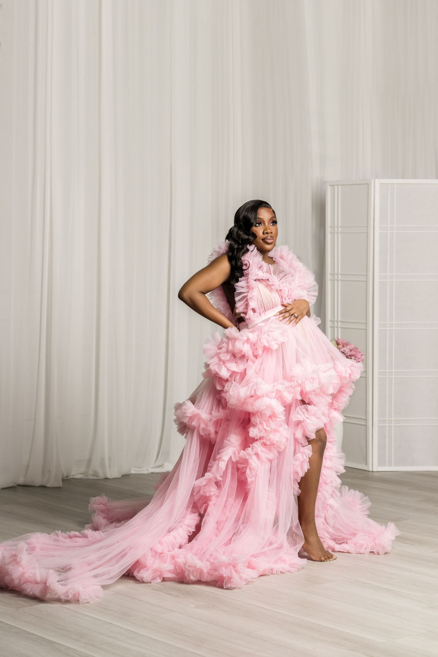 A model poses in a flowing pink tulle gown with dramatic ruffles against a white studio backdrop.
