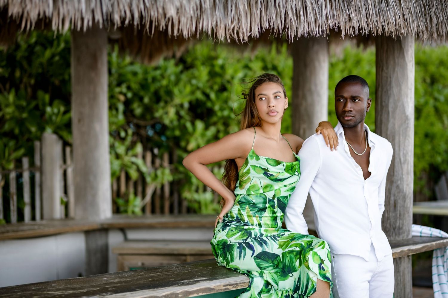 Couple in white and tropical print outfits pose under thatched roof pavilion.