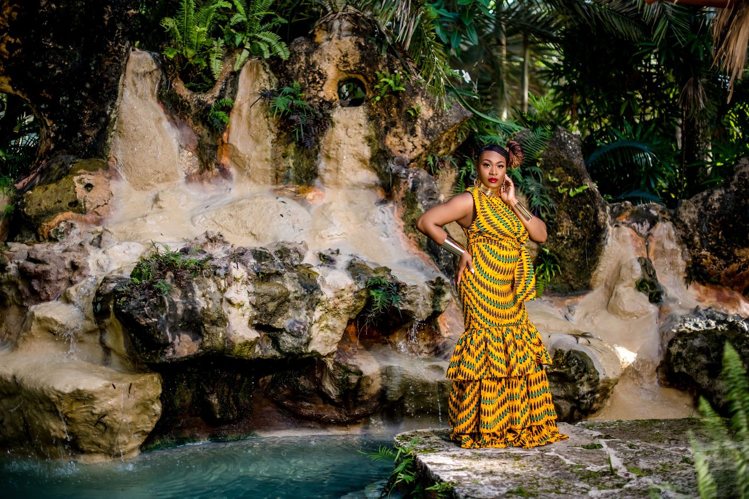 Woman in yellow striped dress poses near a scenic waterfall and rock formations in a tropical setting.