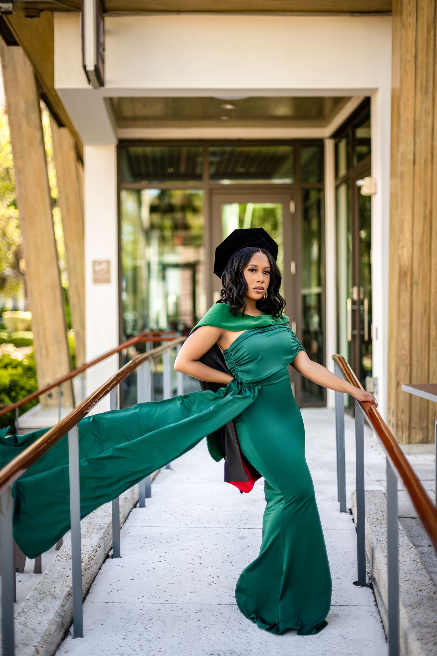 A woman in a flowing emerald green dress poses dramatically on a covered walkway with wooden pillars and handrails.