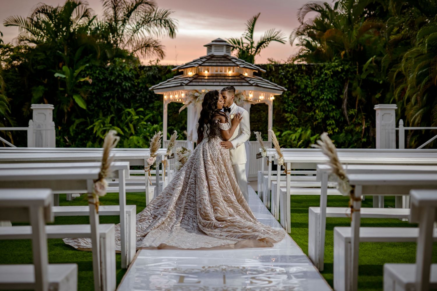 Garden wedding ceremony setup with white gazebo, chairs and palm trees during sunset.