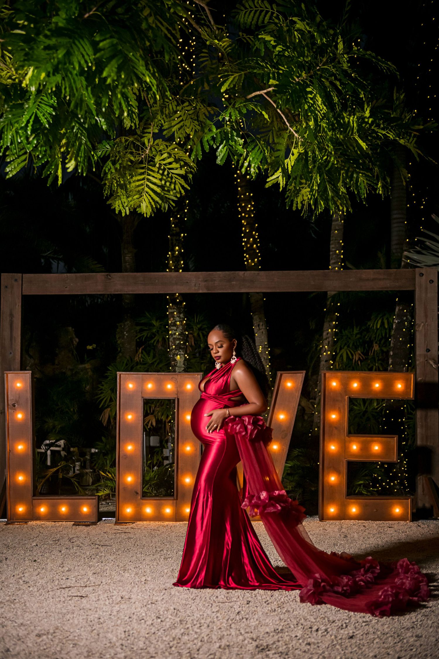 A figure in a flowing red gown poses elegantly by illuminated marquee letters spelling LOVE in an outdoor garden setting.