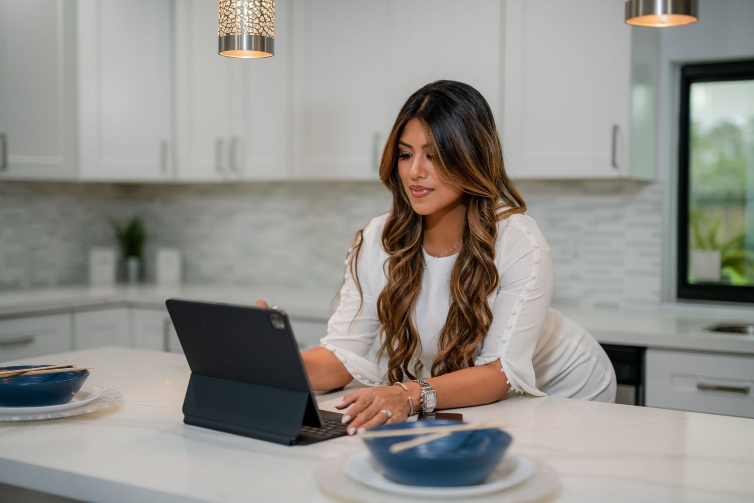 Woman working from home on tablet in modern white kitchen interior.