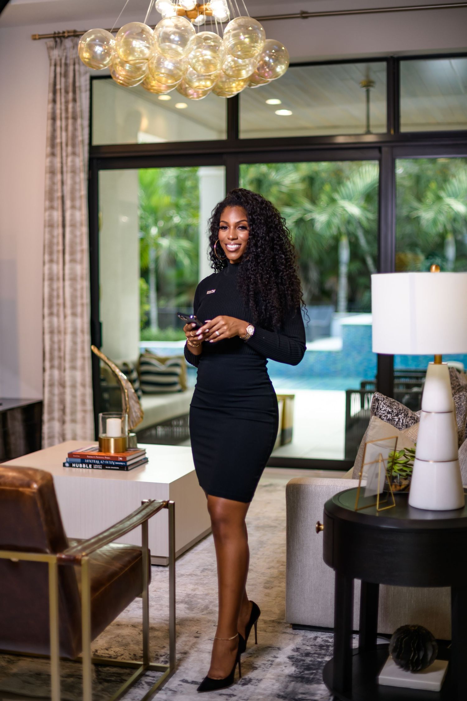 Professional woman in black dress standing in modern luxury interior with pool view.