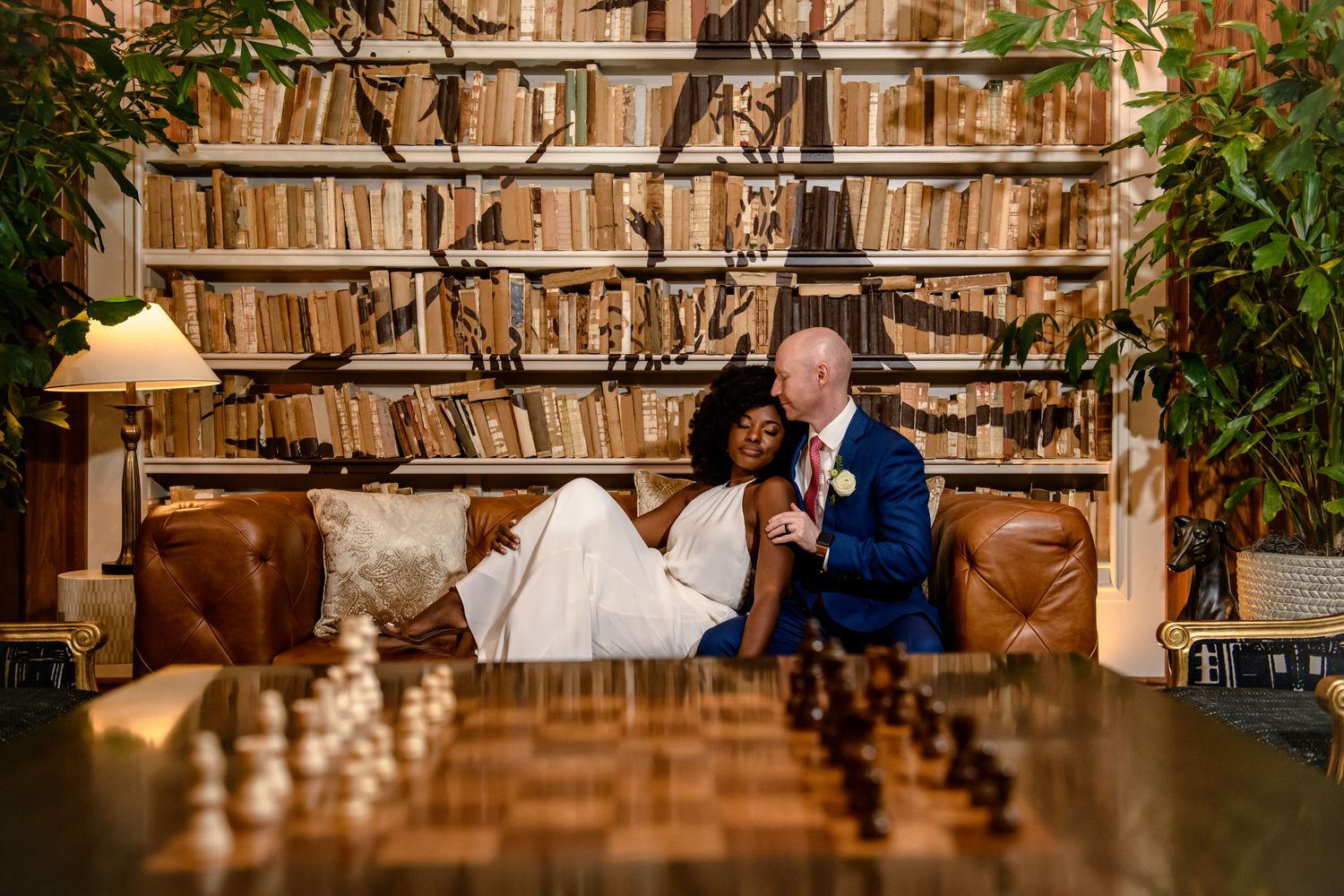 Two people sit at a rustic bar counter with wood paneling and shelving displaying bottles in warm lighting.