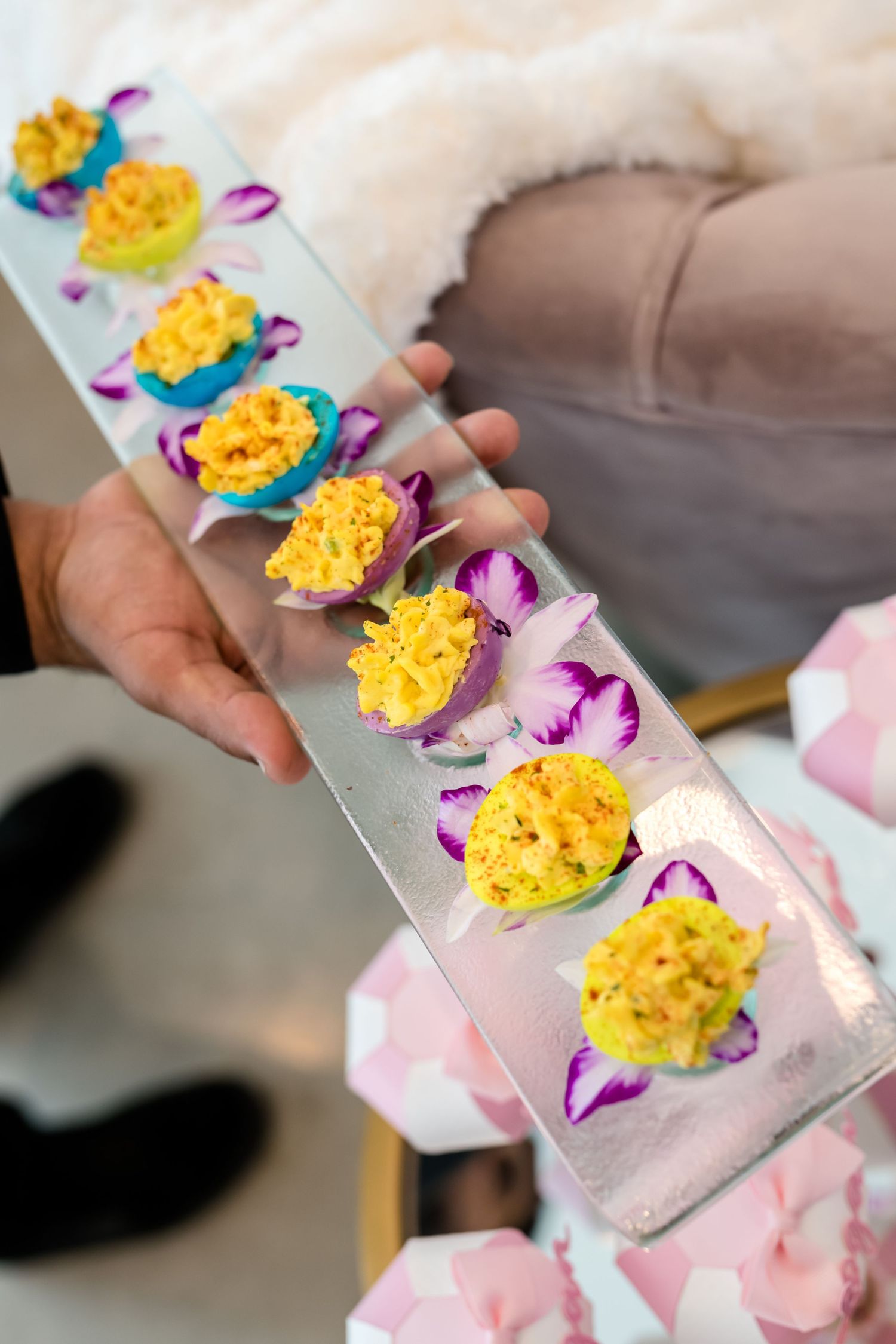 A hand holds a long white serving plate with yellow appetizers garnished with purple flower petals.