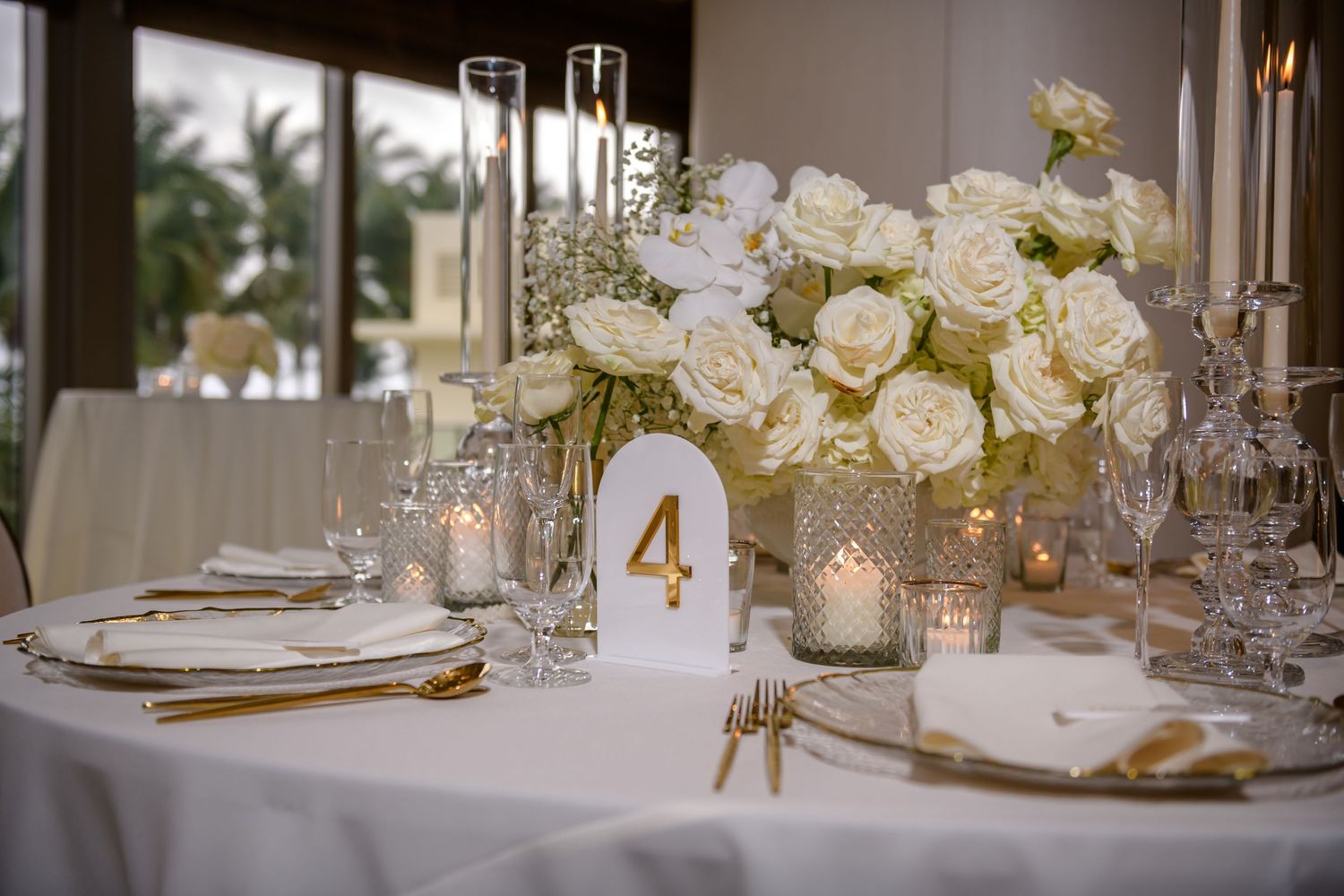 Elegant wedding reception tables decorated with white roses, gold accents, and glowing candles in a luxurious ballroom setting.