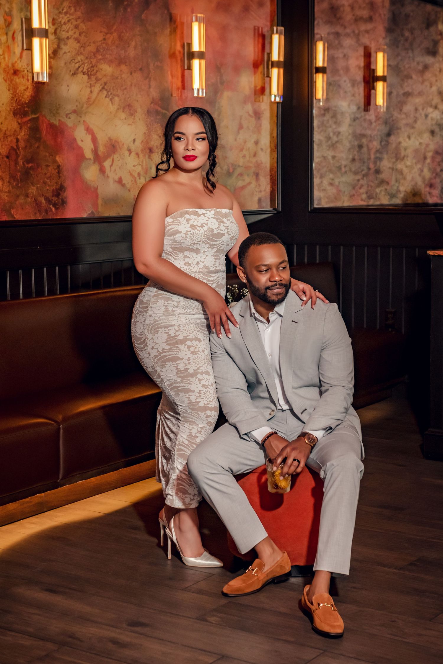 Elegant couple posing in upscale restaurant, woman in white lace dress, man in gray suit seated on ottoman.