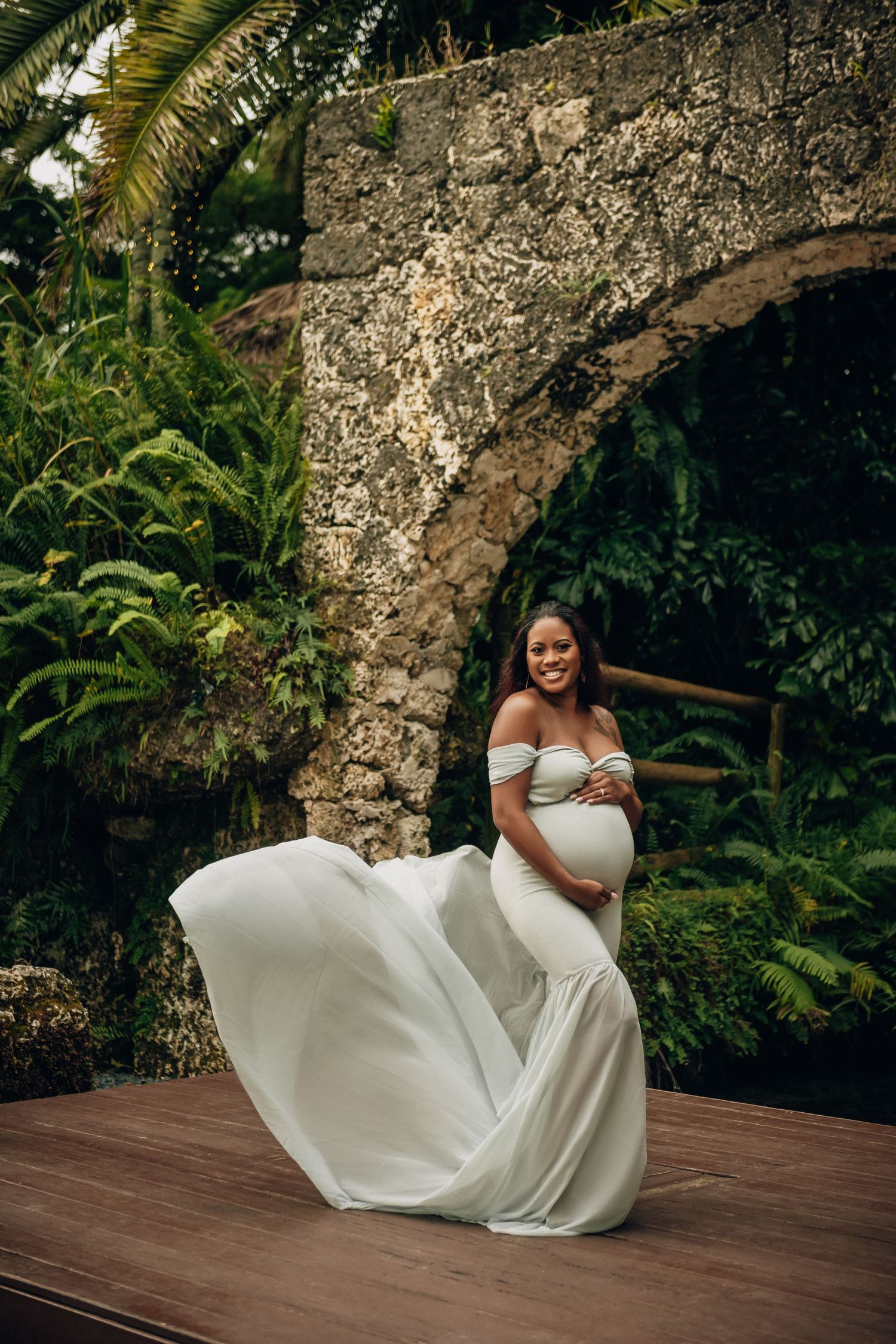 Elegant maternity portrait on wooden deck with stone arch and tropical greenery.