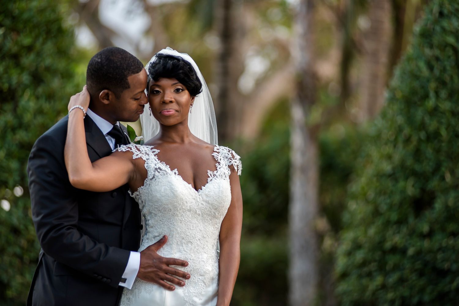 A tender moment between newlyweds outdoors with a dreamy forest backdrop, bride wearing lace gown.