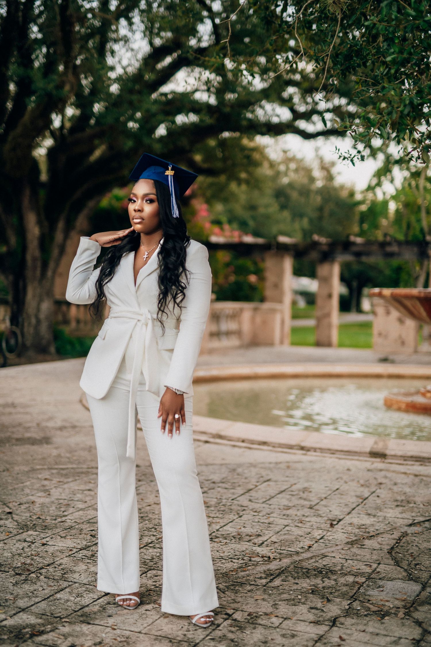 A graduate in a white pantsuit and cap poses outdoors on stone pavement near trees during a graduation photoshoot.