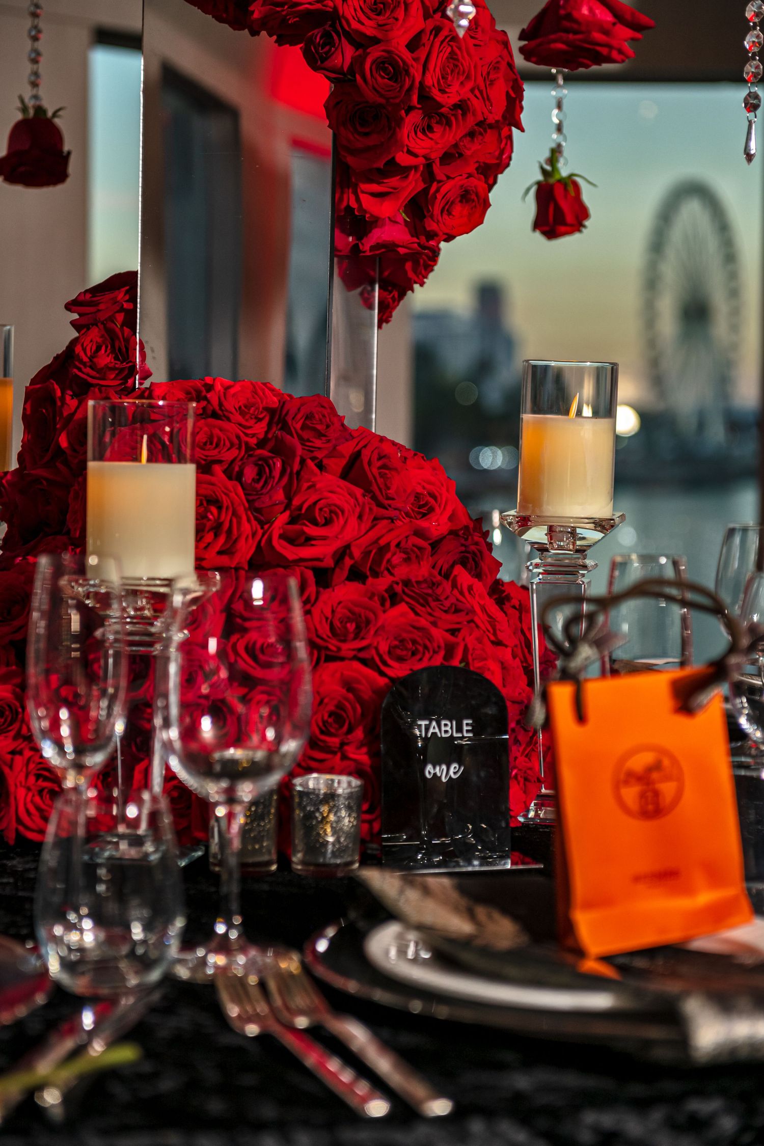 Elegant table setting with red roses and candles against a dark backdrop with city views through windows.