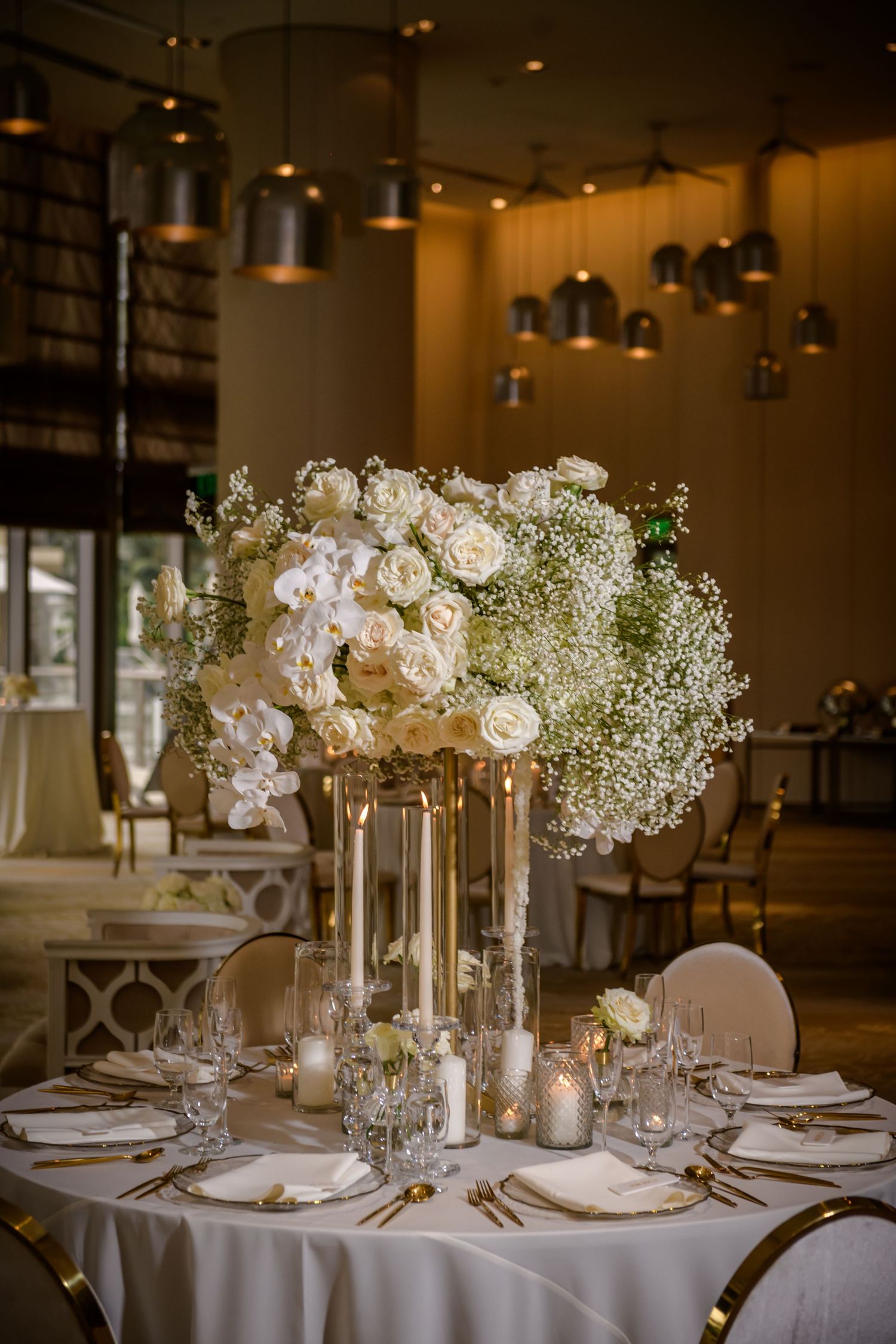 Elegant wedding reception table with white floral centerpiece, candles and crystal glassware in warm-lit venue.