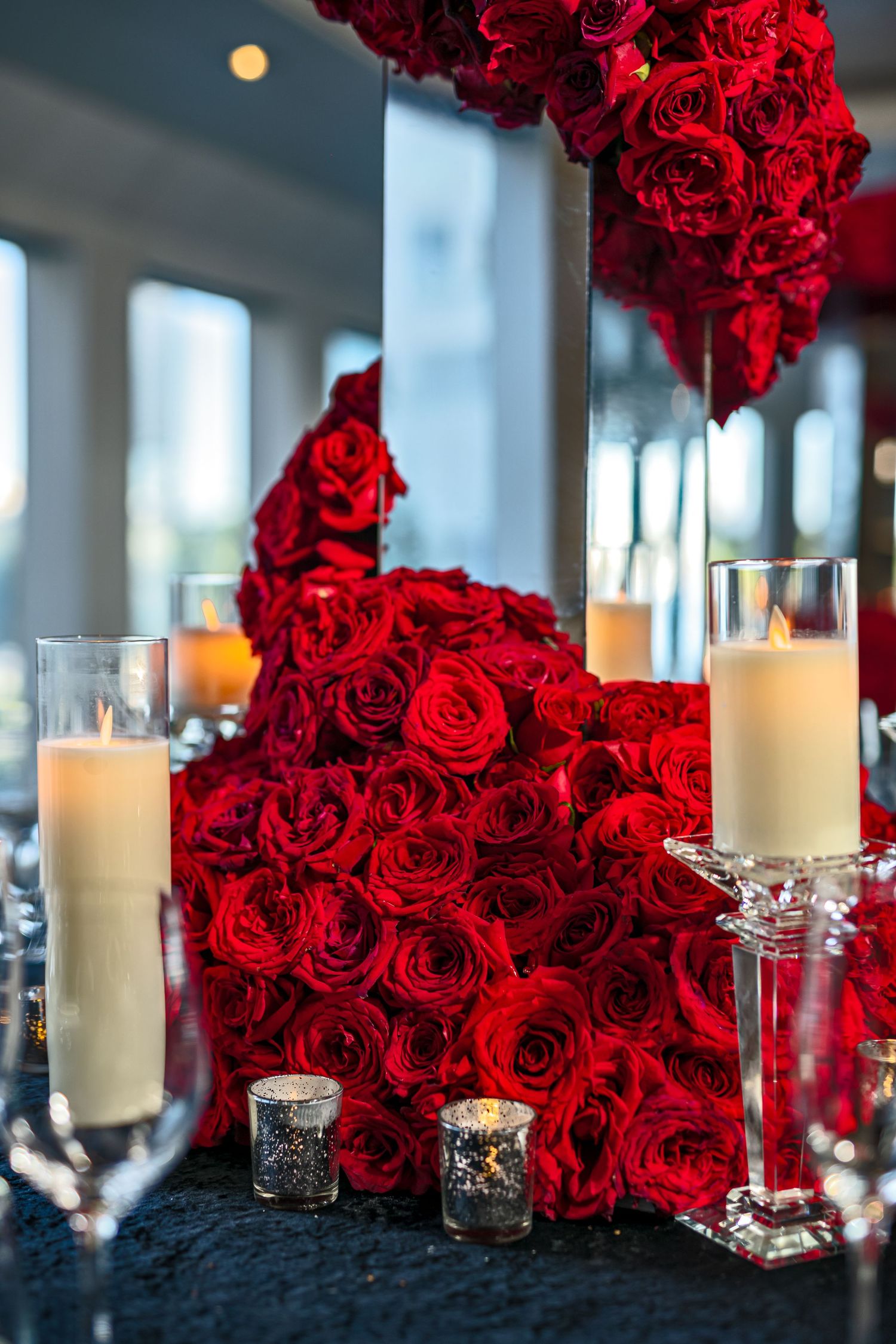 Luxurious wedding reception decor featuring deep red roses and white pillar candles on dark tablecloths.