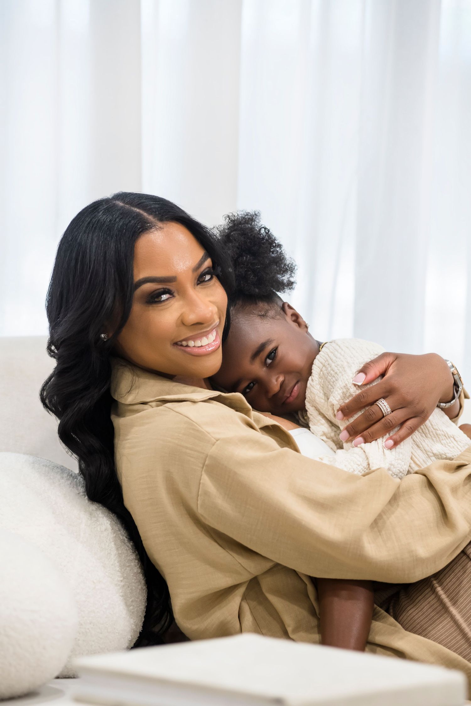 Mother and child sharing a tender embrace while relaxing on white couch in beige loungewear.
