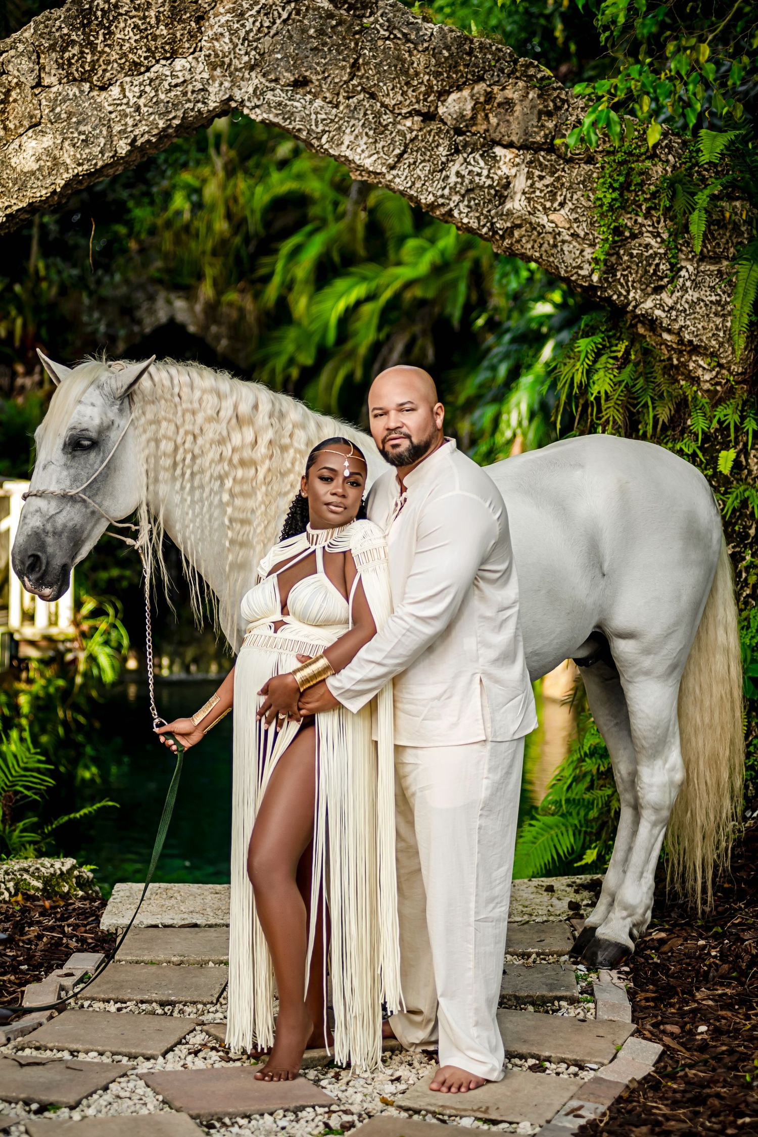 Elegant couple in white outfits pose with majestic white horse in tropical garden setting.