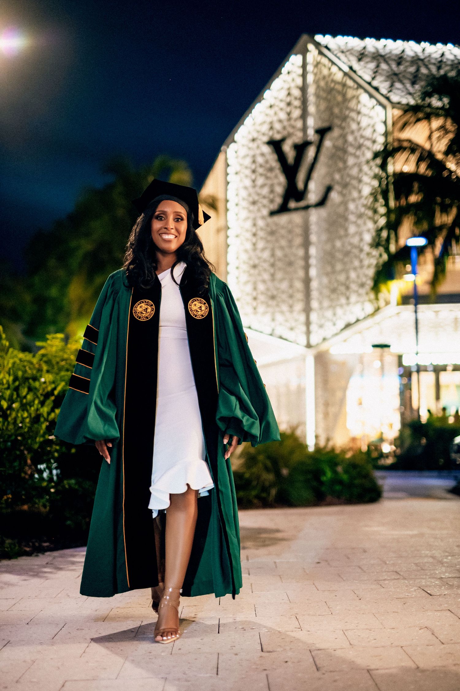 A graduate in green academic regalia poses for outdoor graduation photos at night with illuminated architecture.