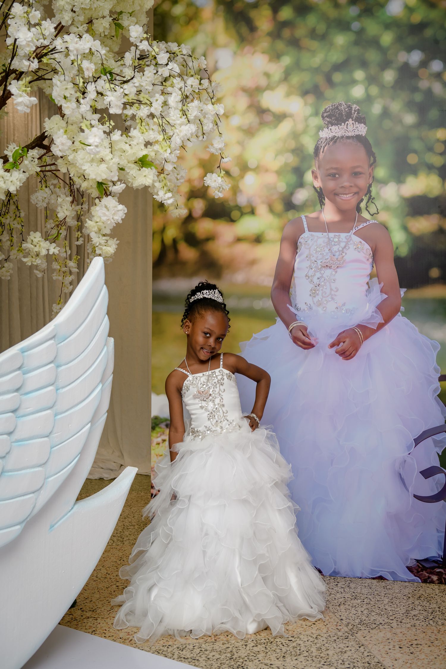 Two children in white formal dresses sit together outdoors under flowering trees for a special occasion photo shoot.