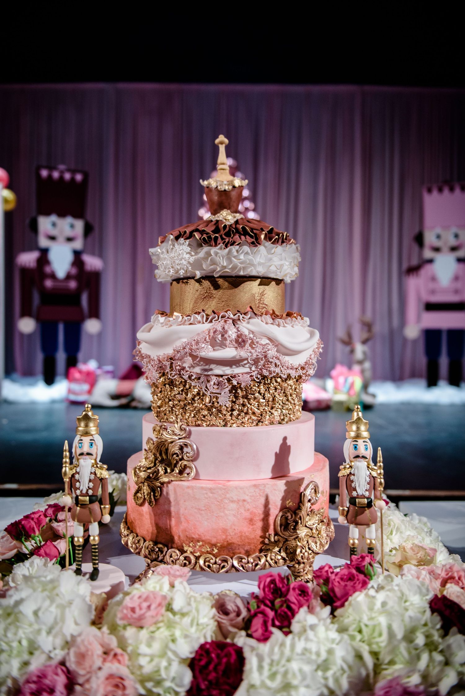 Multi-tiered pink cake decorated with gold trim and robot figurines surrounded by pink and white flowers.