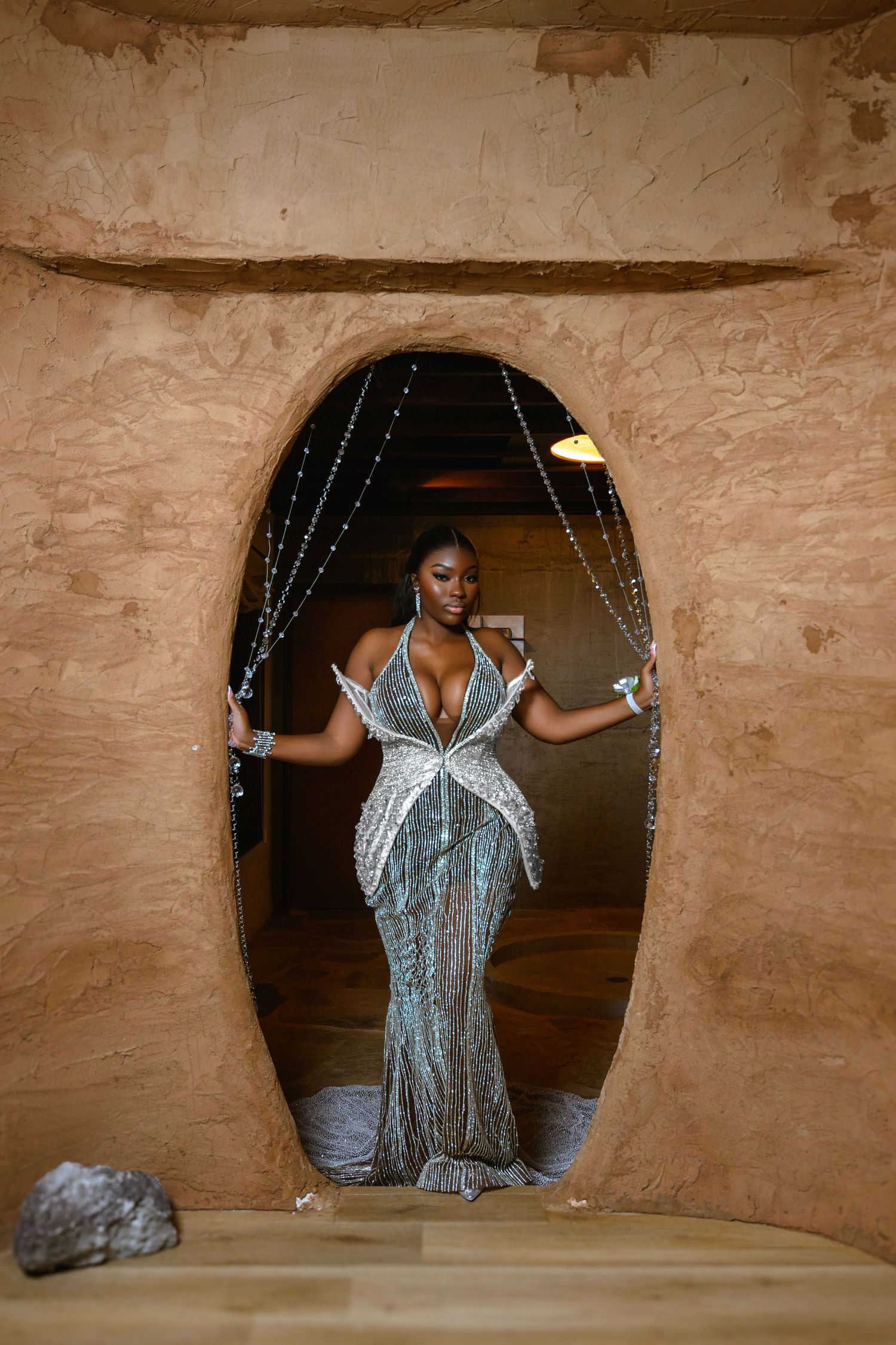 Woman in silver sequin gown posing in oval doorway with beaded curtains in earthy textured setting.