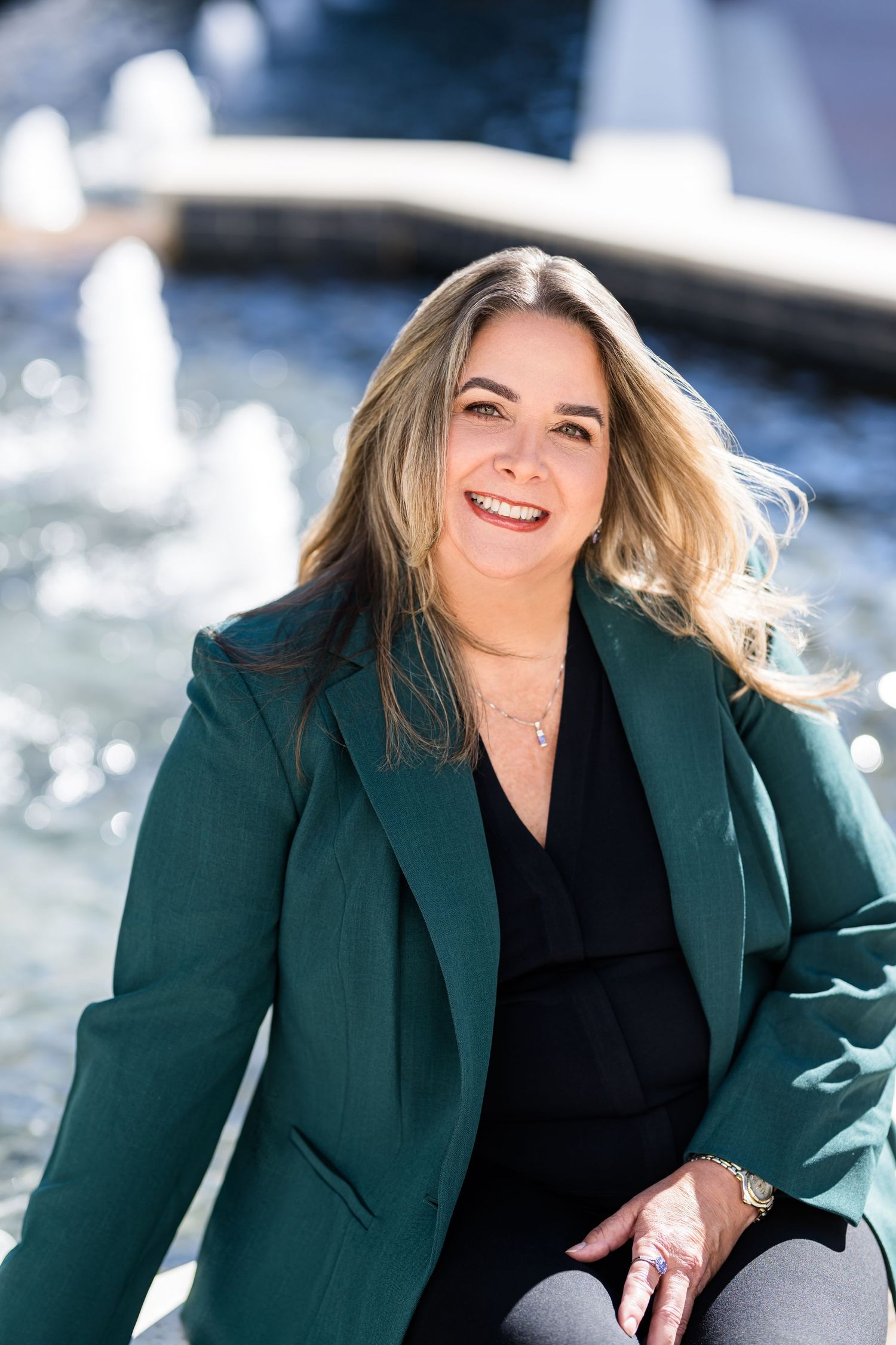 A woman in a green blazer and black top poses outdoors with a bright smile against a snowy winter background.