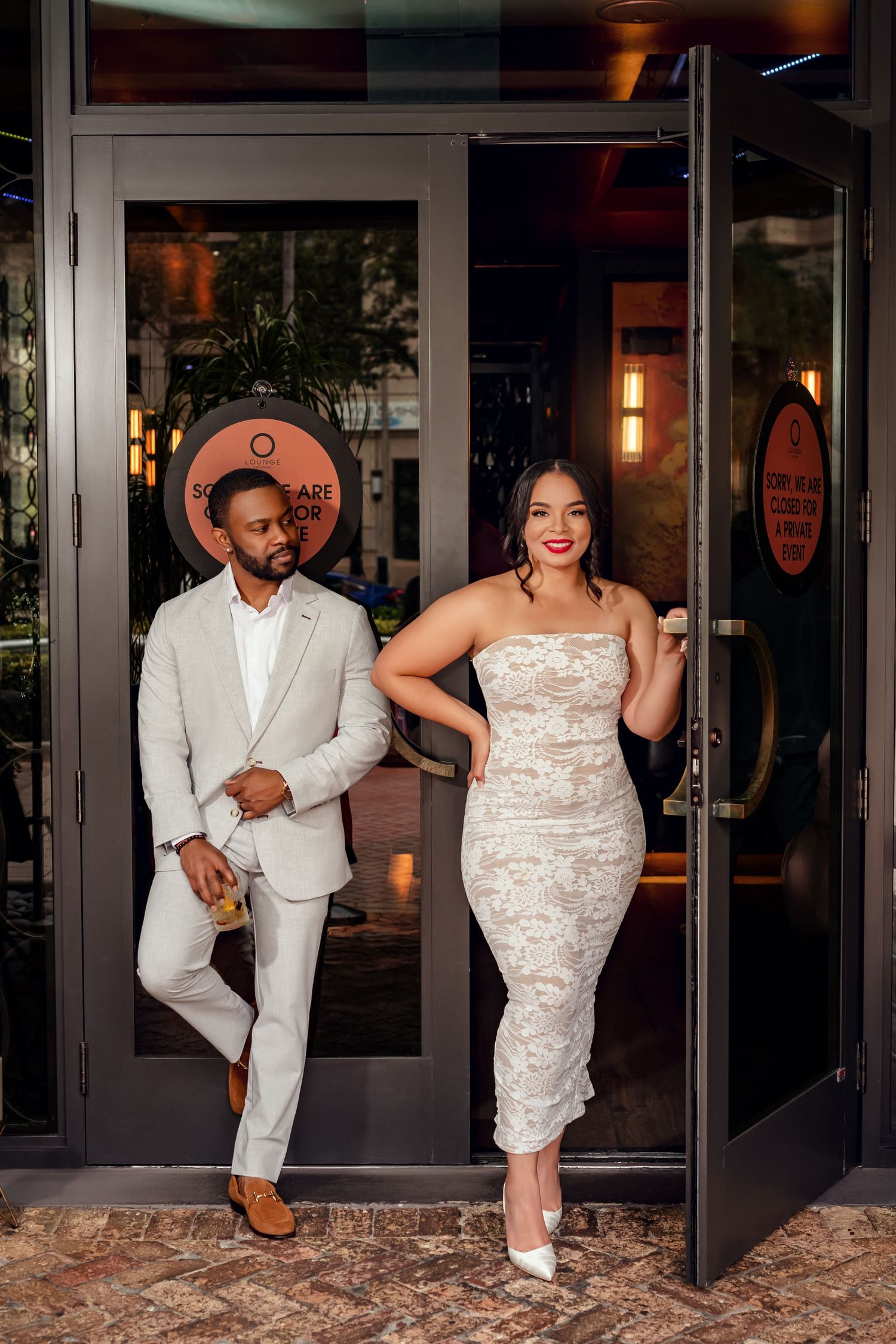 Elegant couple posing at restaurant entrance, man in beige suit and woman in white lace strapless dress with heels.