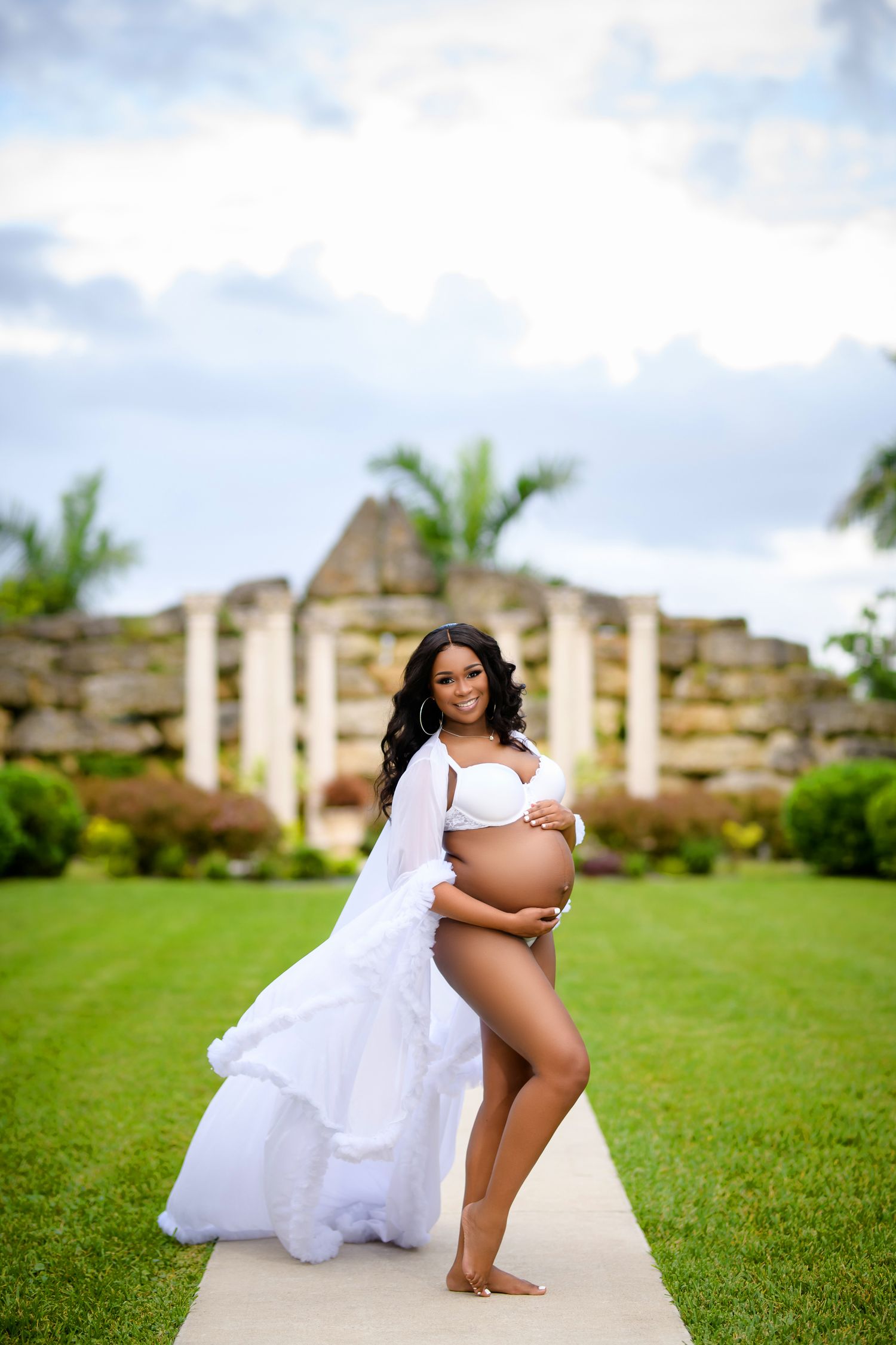 A maternity photoshoot in a white flowing dress on a grassy lawn with stone buildings in the background.