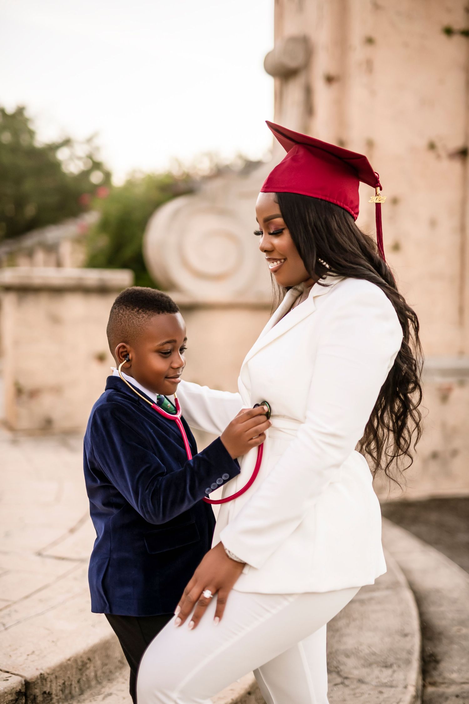 A young child in navy graduation attire hugs someone in white during an outdoor graduation ceremony.