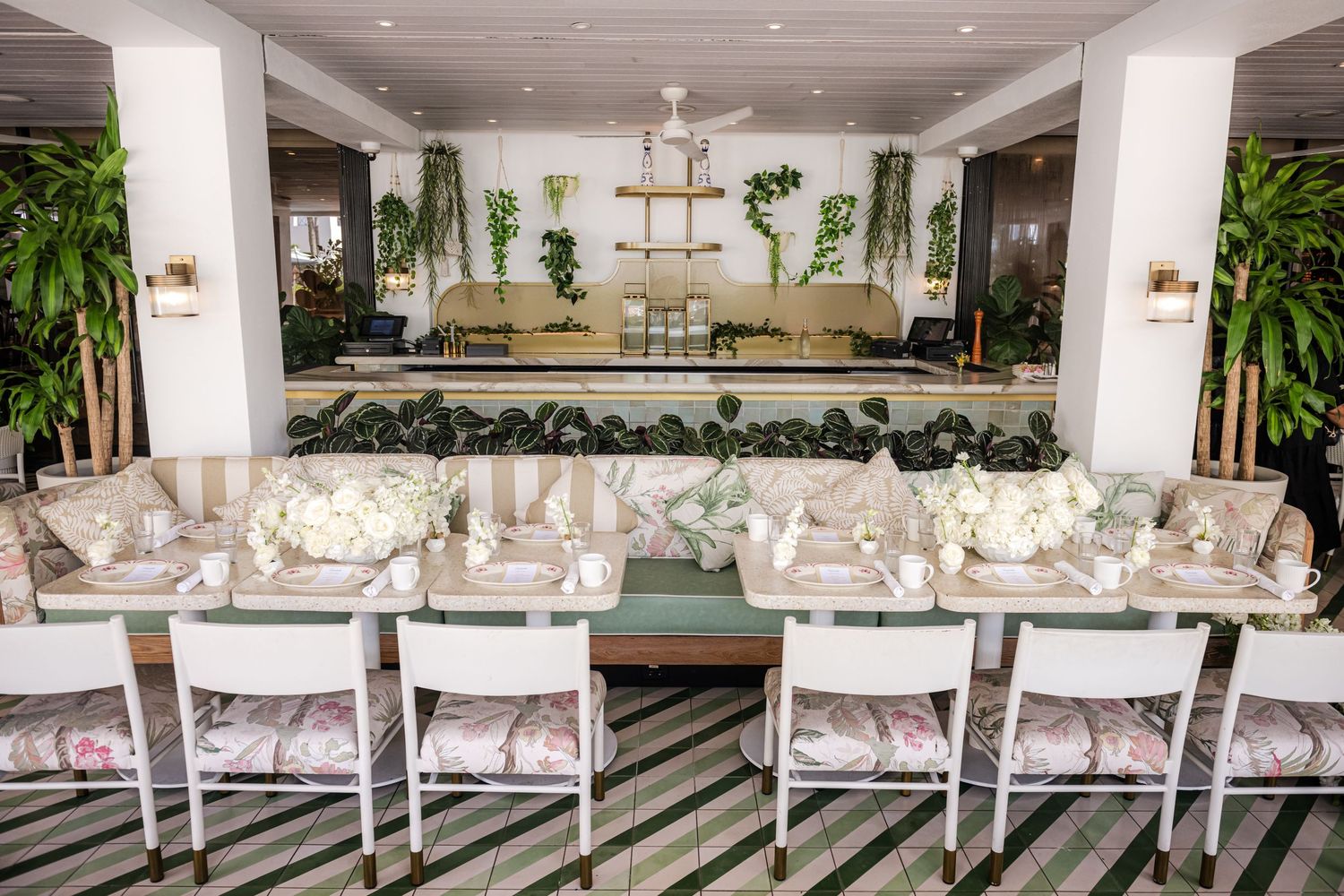 Elegant white dining room with long tables, upholstered chairs, and hanging plants creating a bright, modern atmosphere.