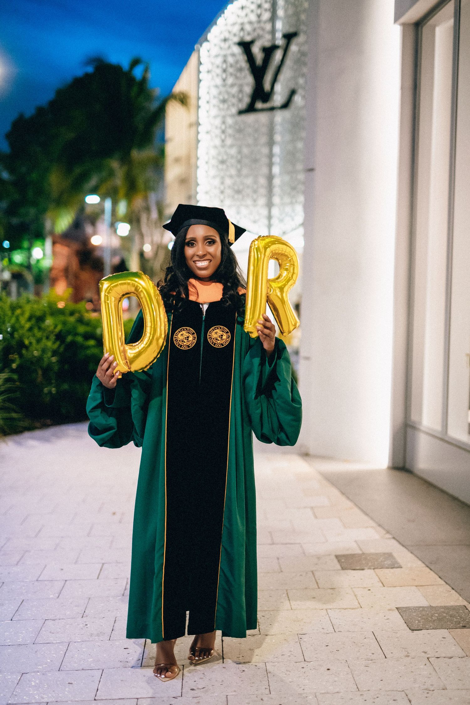 Graduate in emerald green academic regalia holds gold letter balloons while posing for graduation photos outdoors.
