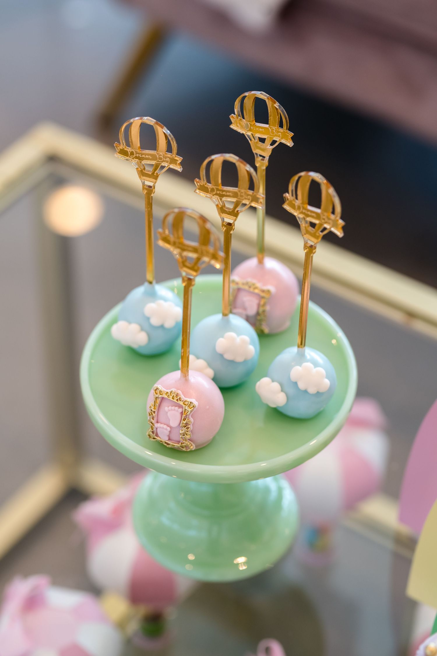 Pastel cake pops decorated with gold crown toppers displayed on a mint green dessert stand.