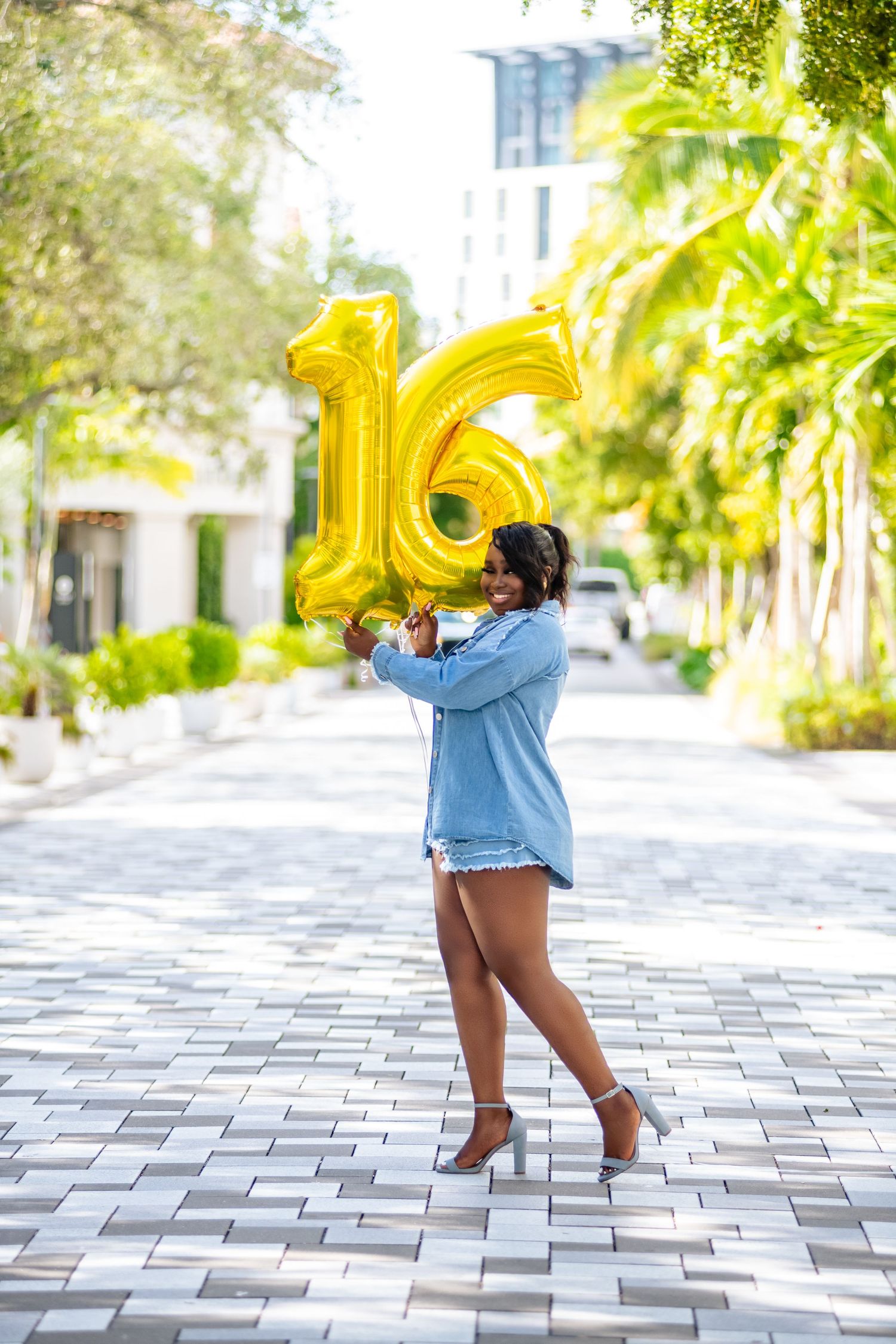 Person in denim shorts holds large gold number 16 balloons while dancing on a sunny outdoor patio.