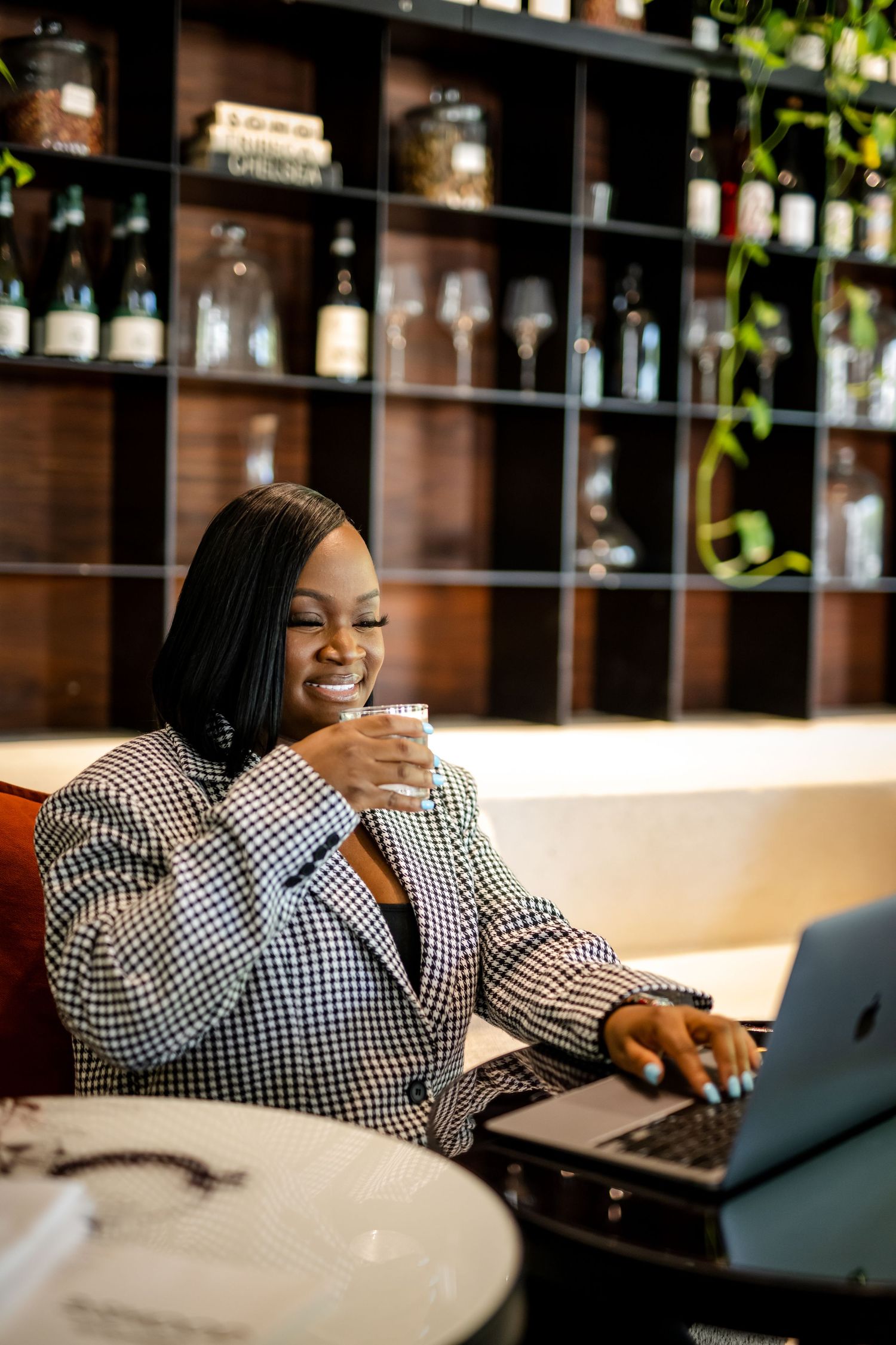 Professional working at laptop in modern office with bottles and shelves in background, wearing checkered blazer.