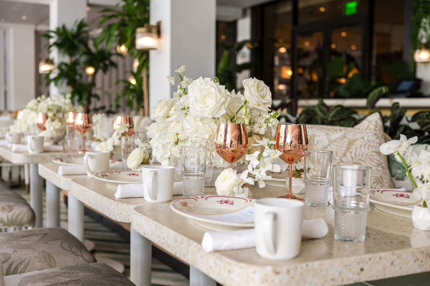 Elegant white and gold wedding reception tables set with floral centerpieces and crystal glassware in a bright venue.