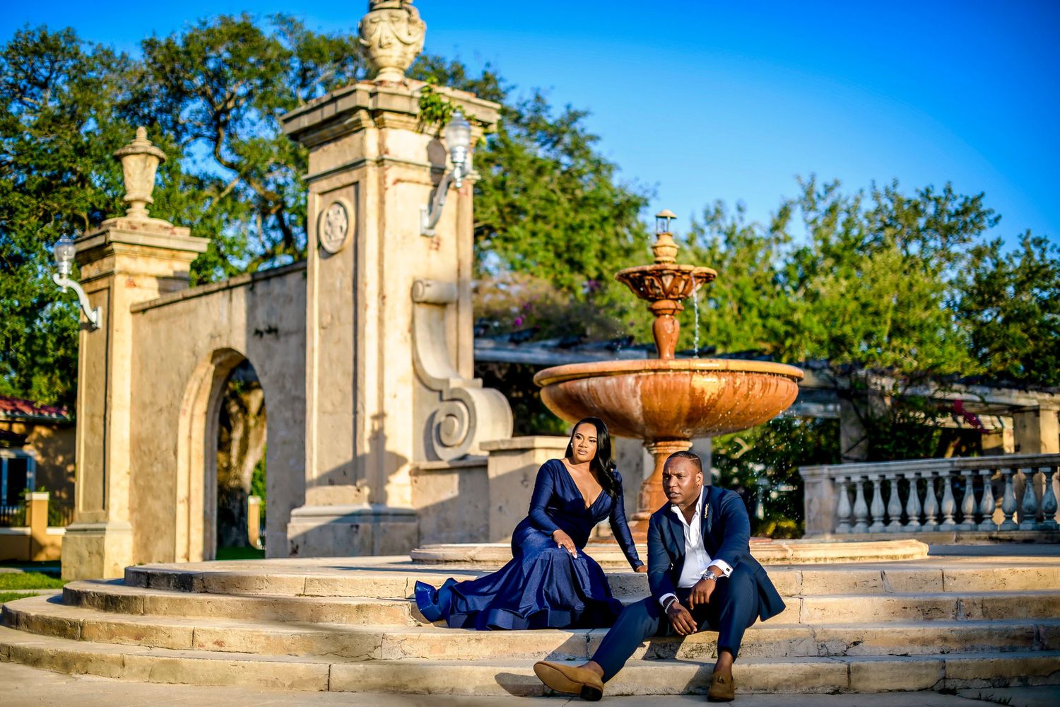Elegant couple in formalwear sits on grand stone steps near an ornate fountain and decorative gate pillars outdoors.