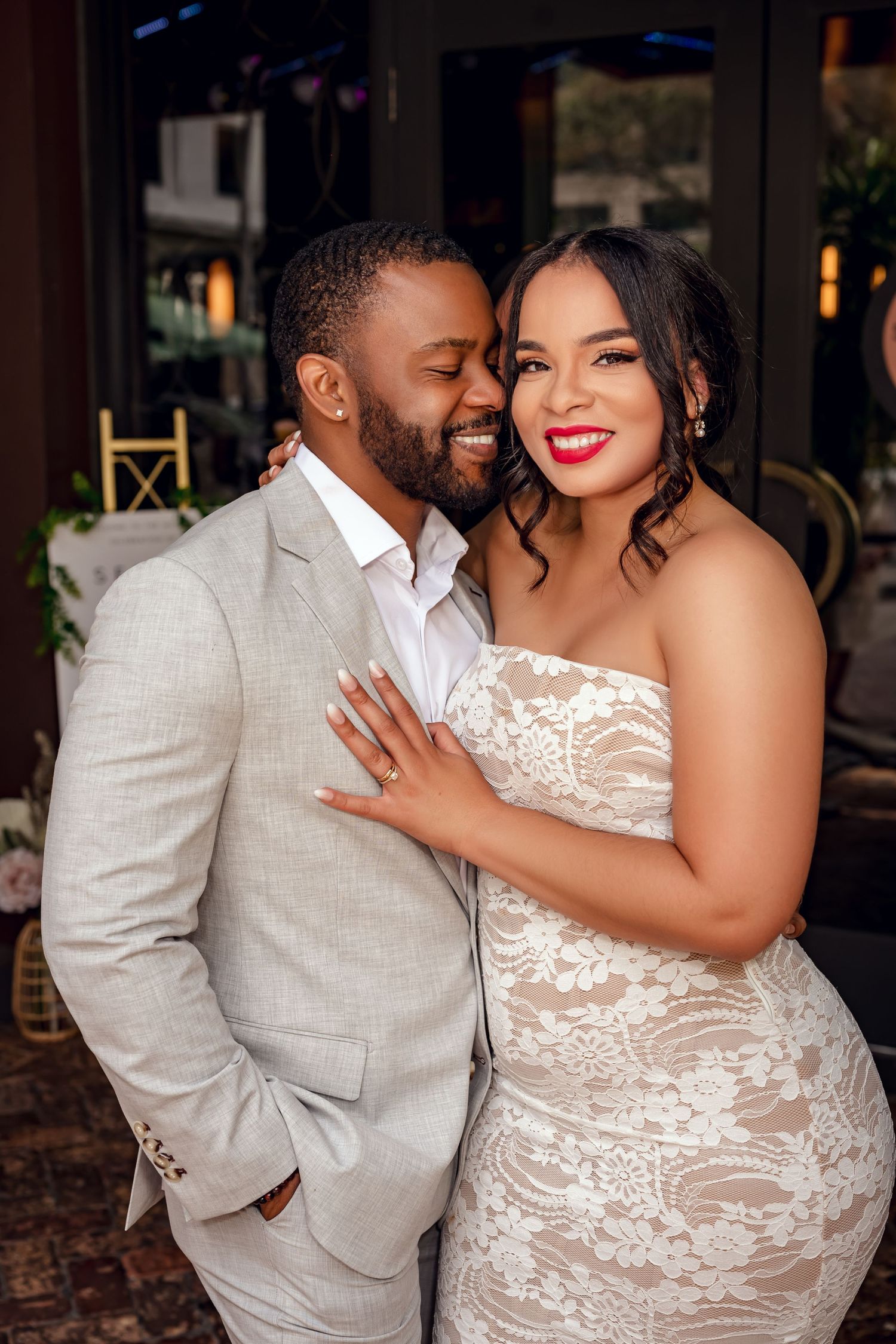 A smiling couple in wedding attire, groom in gray suit and bride in white lace gown, posing together at their reception.